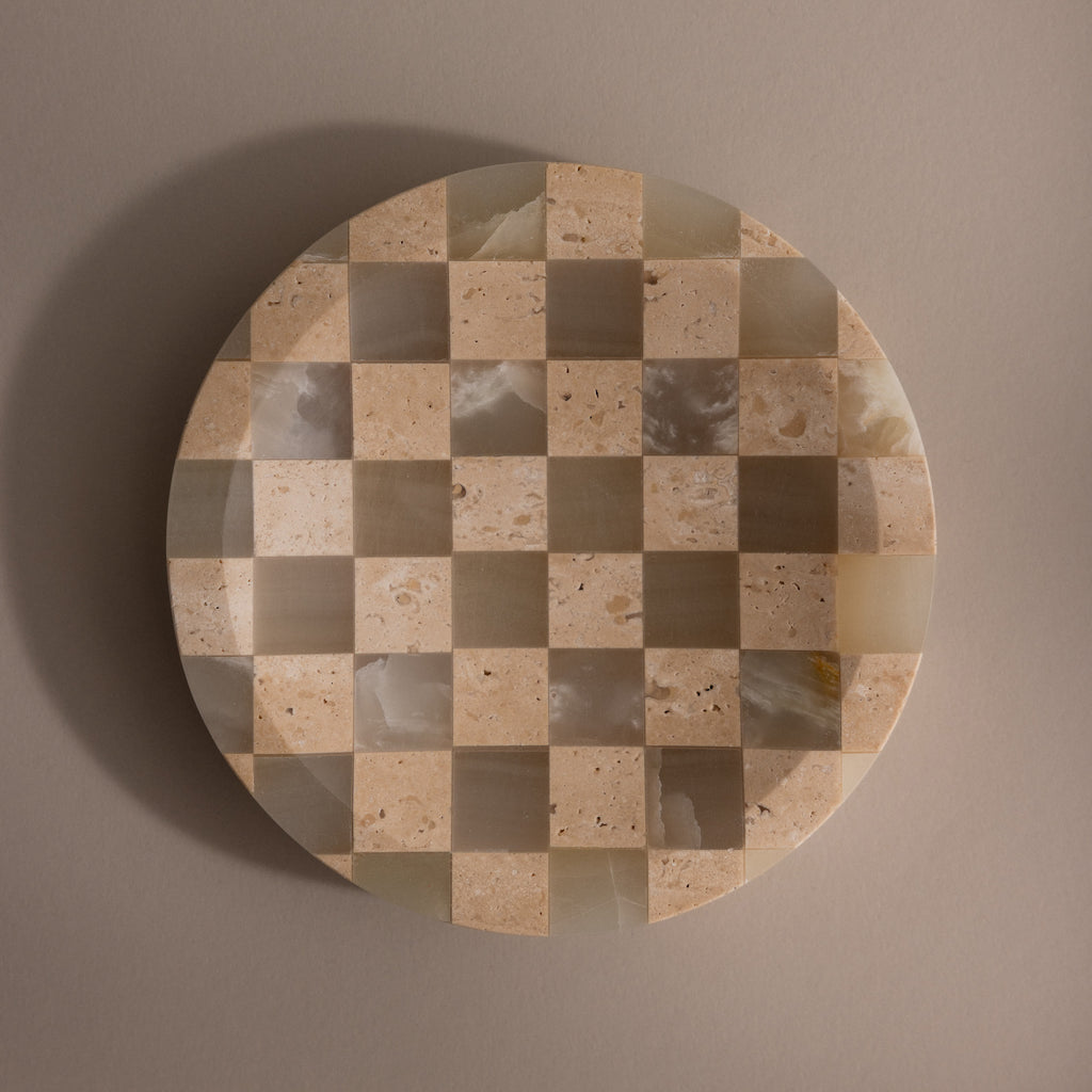 The Checkered Jewelry Dish is a round natural stone tray featuring a beige and cream checkerboard pattern, ideal for displaying jewelry, shown on a plain taupe background.