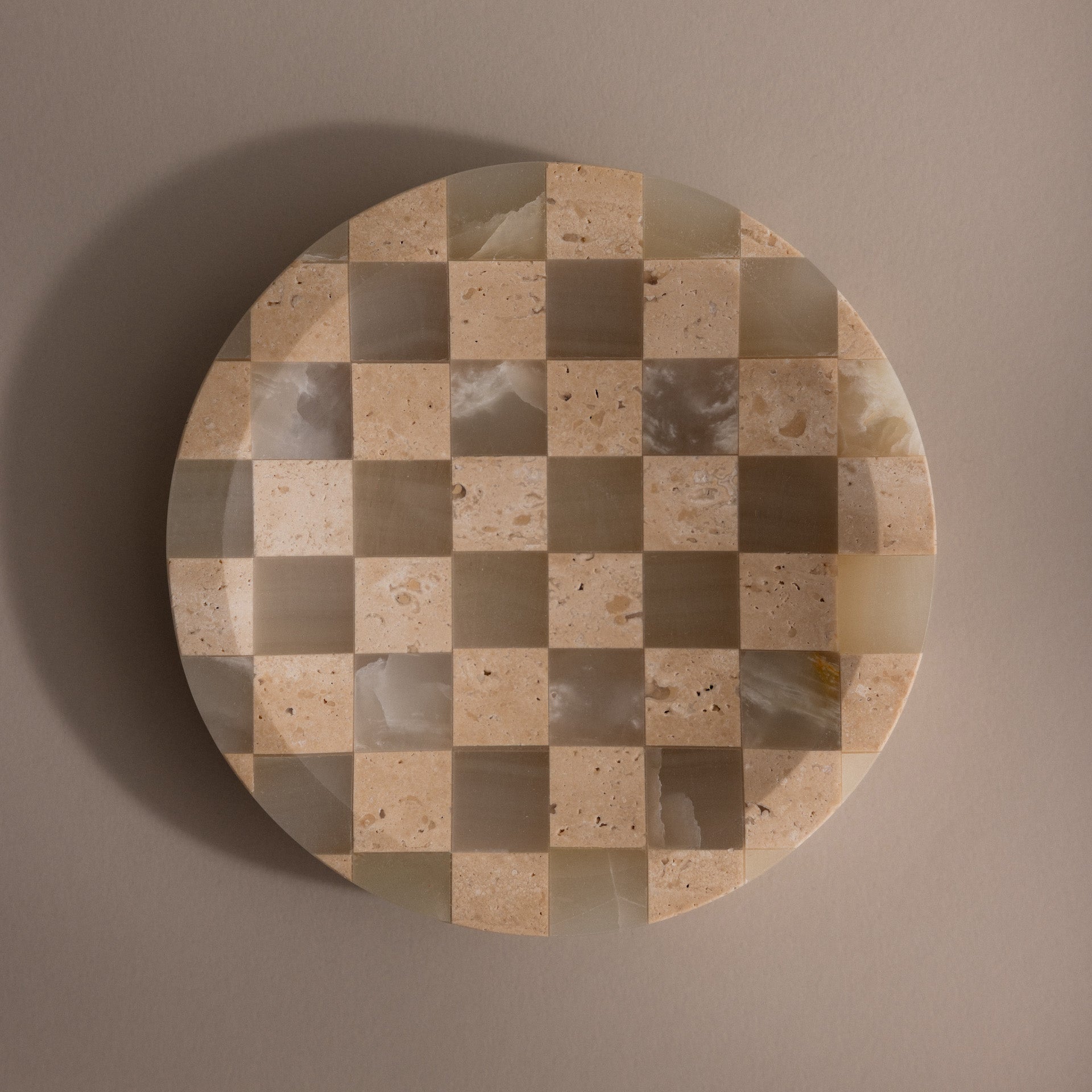 The Checkered Jewelry Dish is a round natural stone tray featuring a beige and cream checkerboard pattern, ideal for displaying jewelry, shown on a plain taupe background.