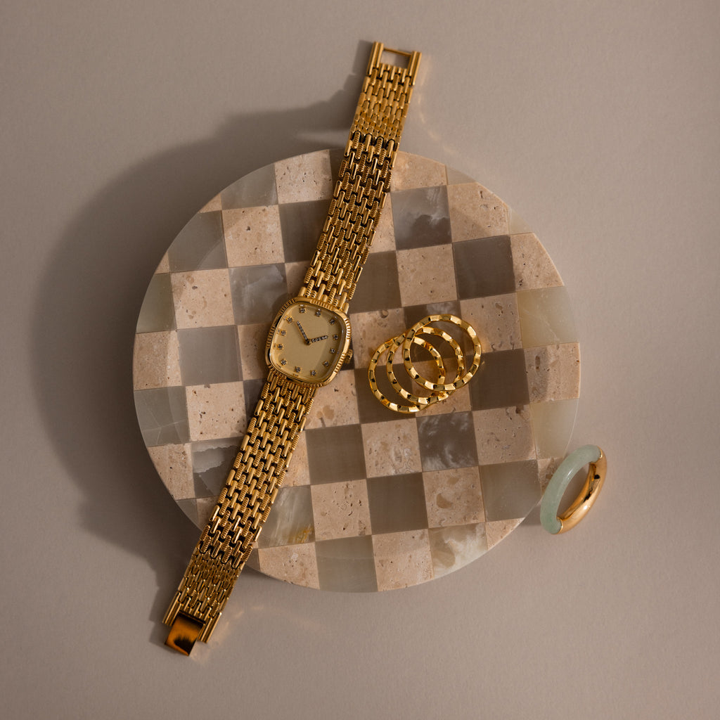 A gold wristwatch, rings, and a jade ring are displayed on the Checkered Jewelry Dish, set against a beige background.