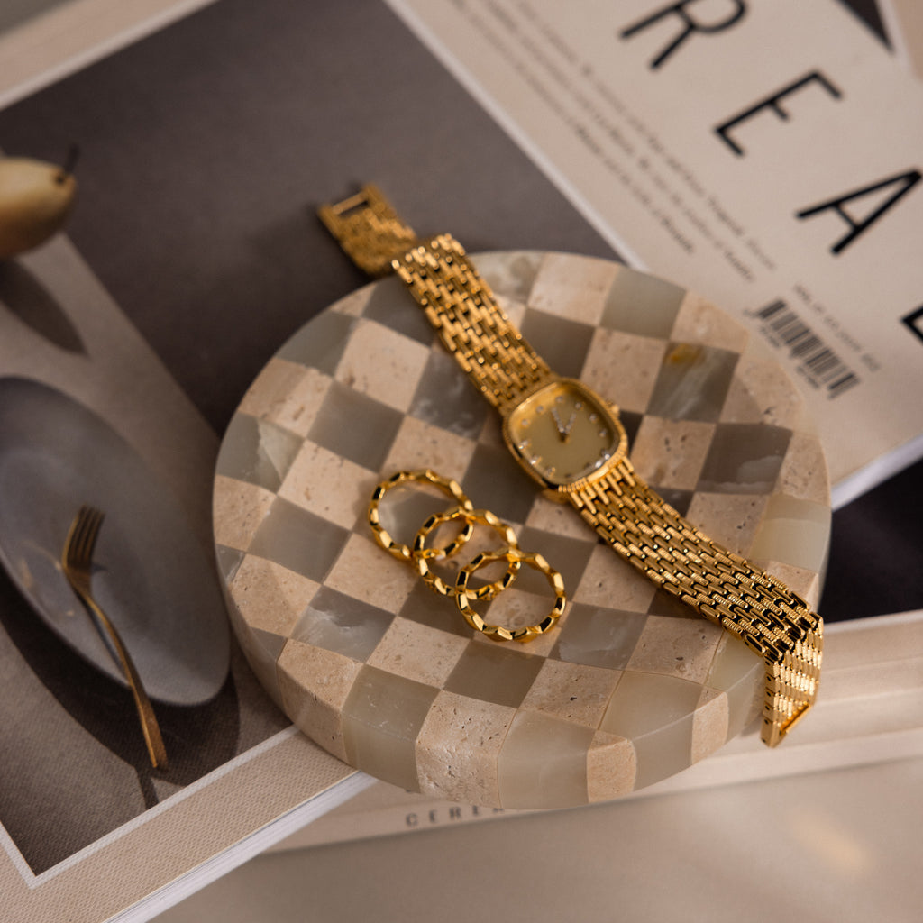 A Checkered Jewelry Dish showcases a gold watch and three gold rings, with magazines and a plate in the background.
