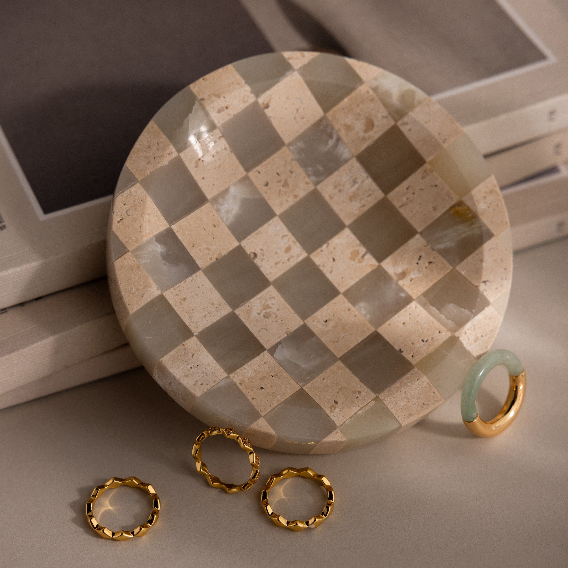 The Checkered Jewelry Dish rests on beige stacked books, stylishly holding three gold rings and one green and gold ring—perfect for blending elegance with everyday function.