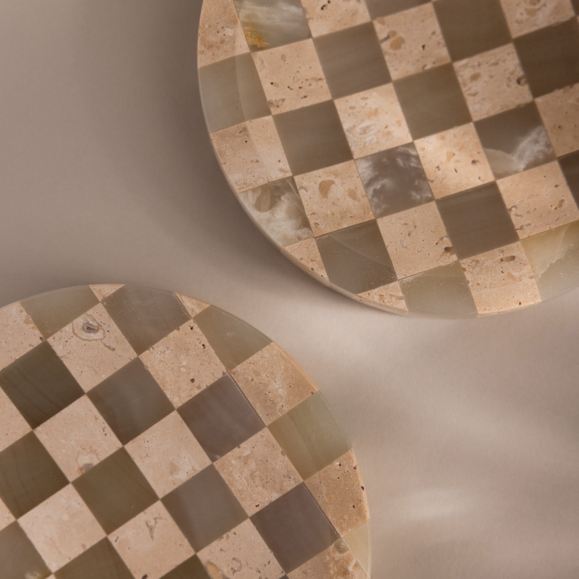 Two round, beige and taupe natural stone dishes with a checkerboard pattern are partially shown on a neutral background—ideal as the Checkered Jewelry Dish to hold your favorite pieces.