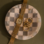 A Gold watch and three gold rings rest on the Checkered Jewelry Dish, a round accessory crafted from natural stone, set against a green backdrop.