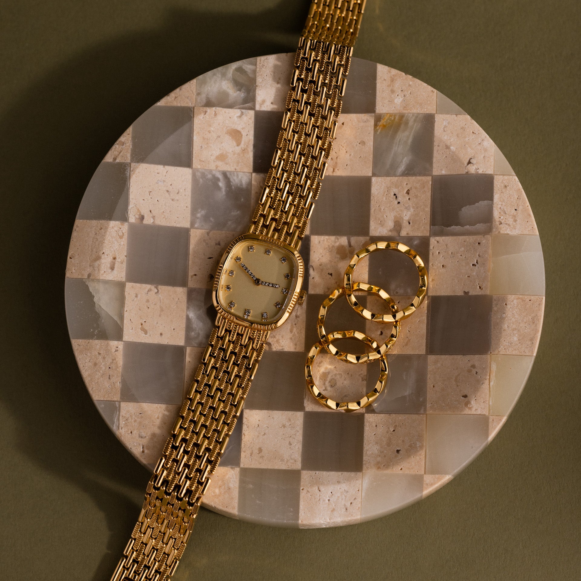 A Gold watch and three gold rings rest on the Checkered Jewelry Dish, a round accessory crafted from natural stone, set against a green backdrop.