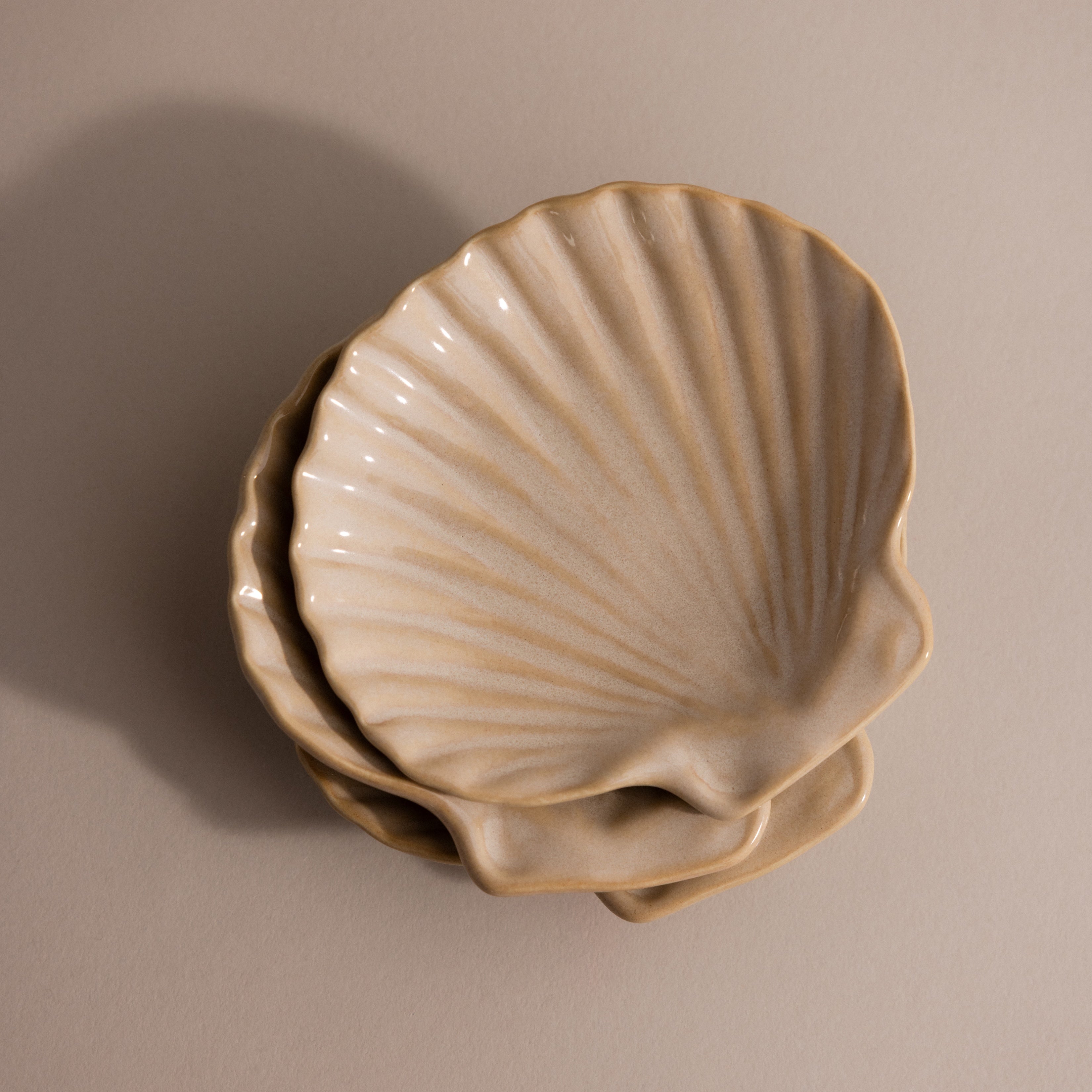 The Venus Shell Jewelry Dish features three beige ceramic plates shaped like seashells, stacked on a neutral background—ideal as a jewelry holder or accent piece for coastal decor.