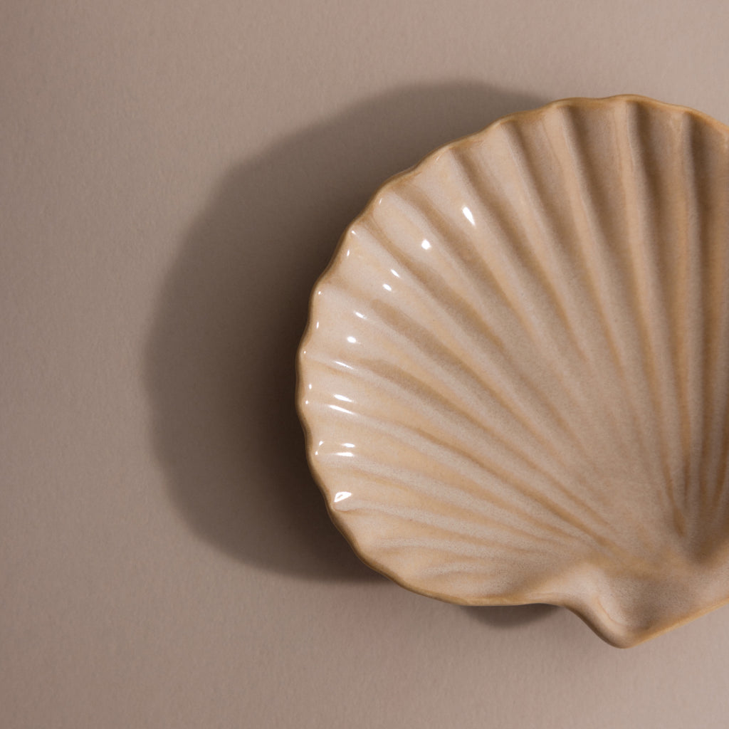 The Venus Shell Jewelry Dish is a beige ceramic scallop shell, ideal as a jewelry holder or accent piece for coastal decor, displayed on a neutral background.