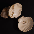 Two beige Venus Shell Jewelry Dishes hold a gold chain and a pearl-accented gold ring on a dark surface—a stylish choice for elegant coastal decor.