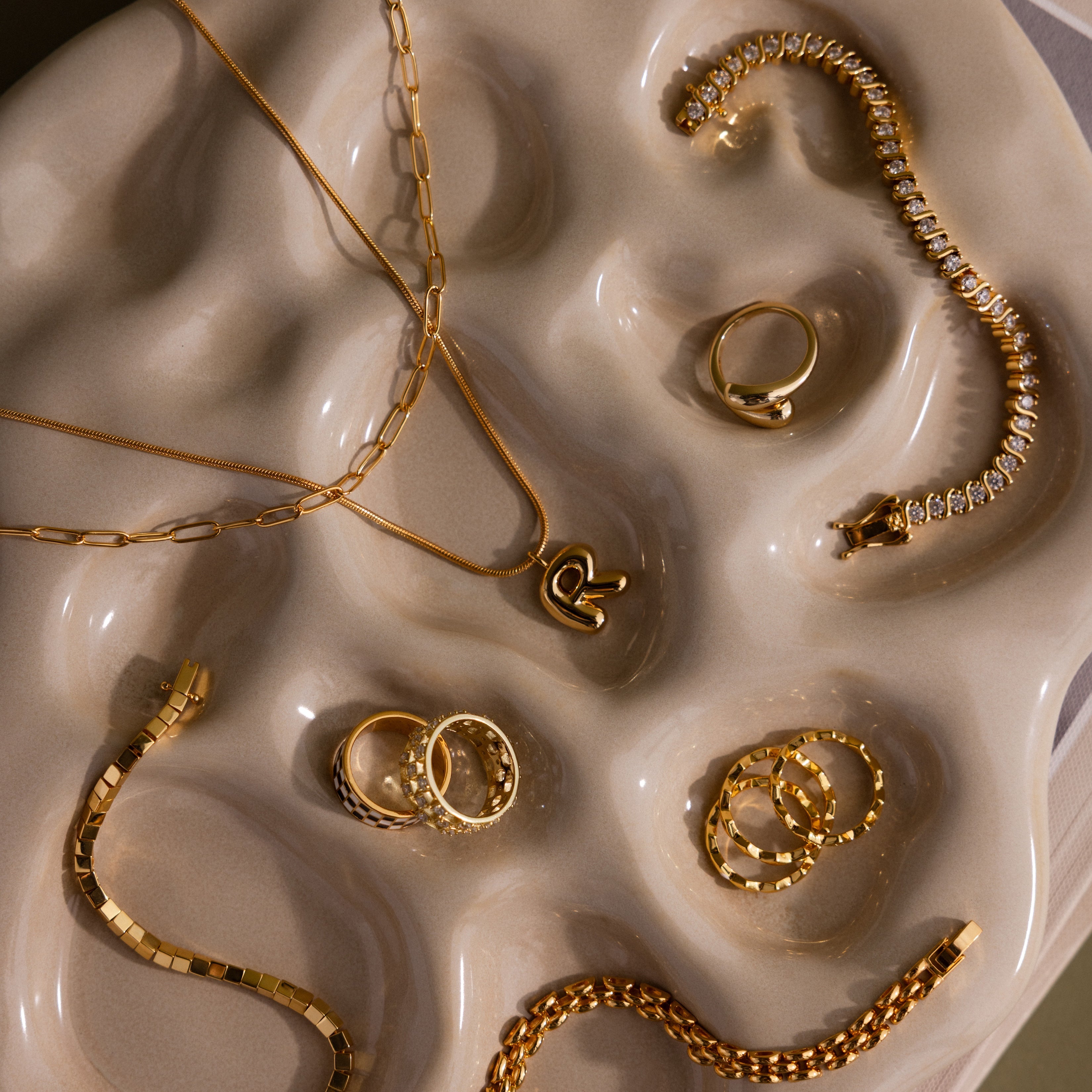 The Organic Treasure Trove Jewelry Dish elegantly displays assorted gold rings, necklaces, and bracelets on its wavy beige minimalist design.