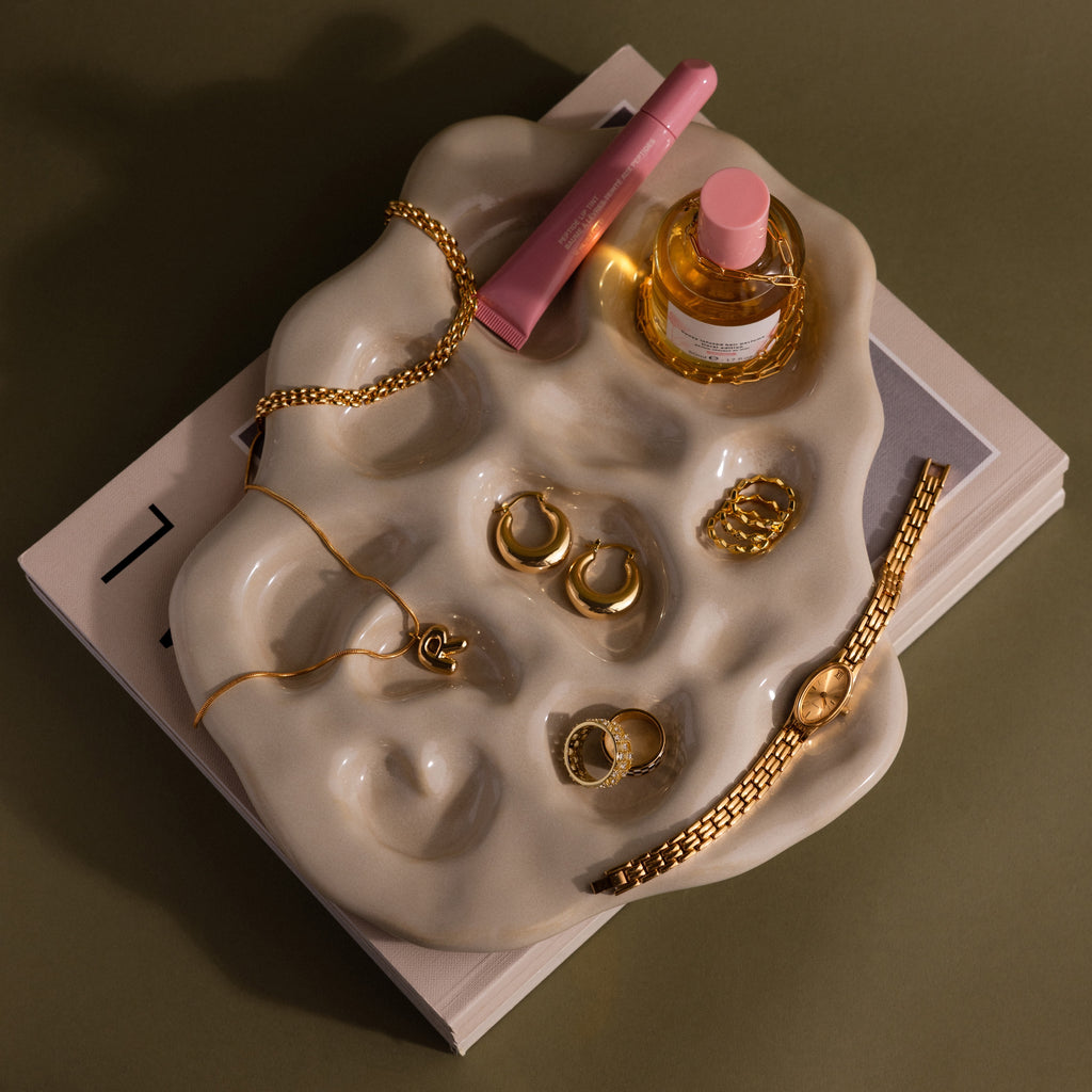The Organic Treasure Trove Jewelry Dish displays gold jewelry, a pink perfume bottle, and lip gloss on stacked books for a chic minimalist look.
