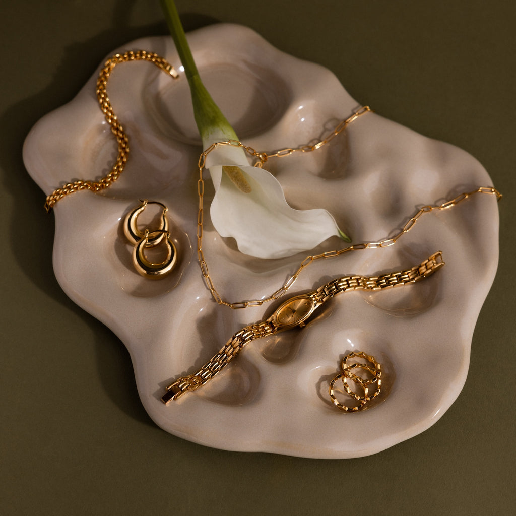The Slim Oval Timepiece in 18K Gold is displayed with gold jewelry and a white flower on an abstract beige ceramic dish, set against an olive green background.