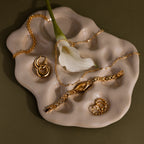 The Slim Oval Timepiece in 18K Gold is displayed with gold jewelry and a white flower on an abstract beige ceramic dish, set against an olive green background.