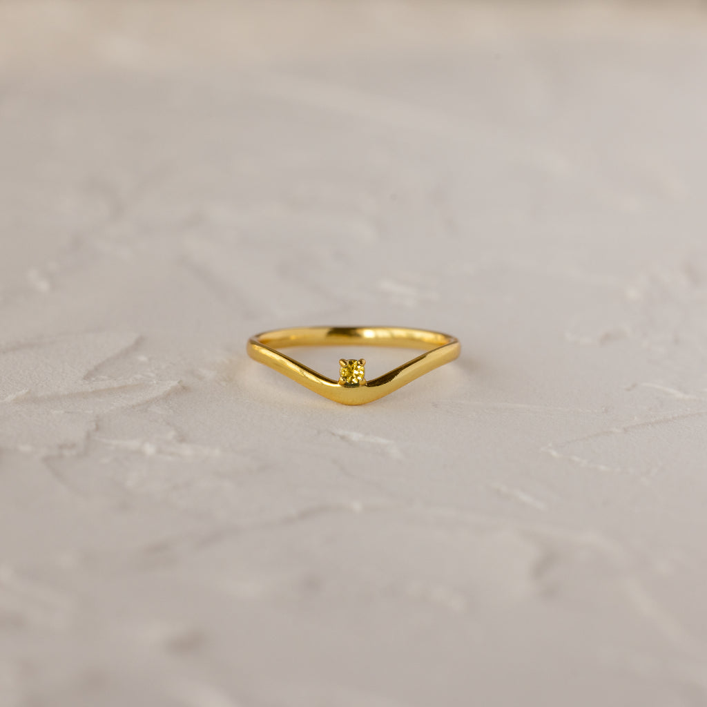 The Topaz Drop Curve Ring, a delicate gold stacking ring, features a single small diamond and is displayed on a textured light-colored surface.