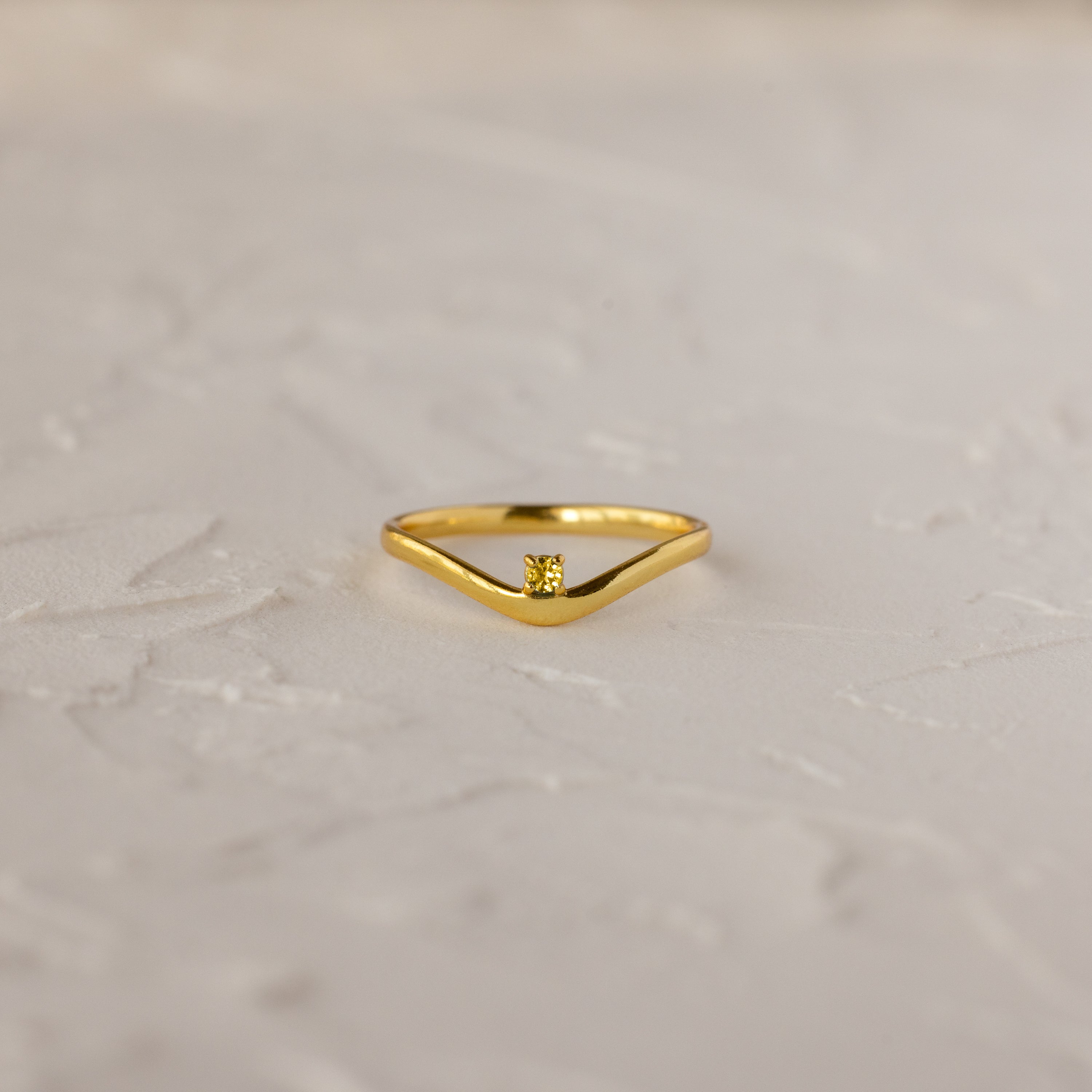 The Topaz Drop Curve Ring, a delicate gold stacking ring, features a single small diamond and is displayed on a textured light-colored surface.