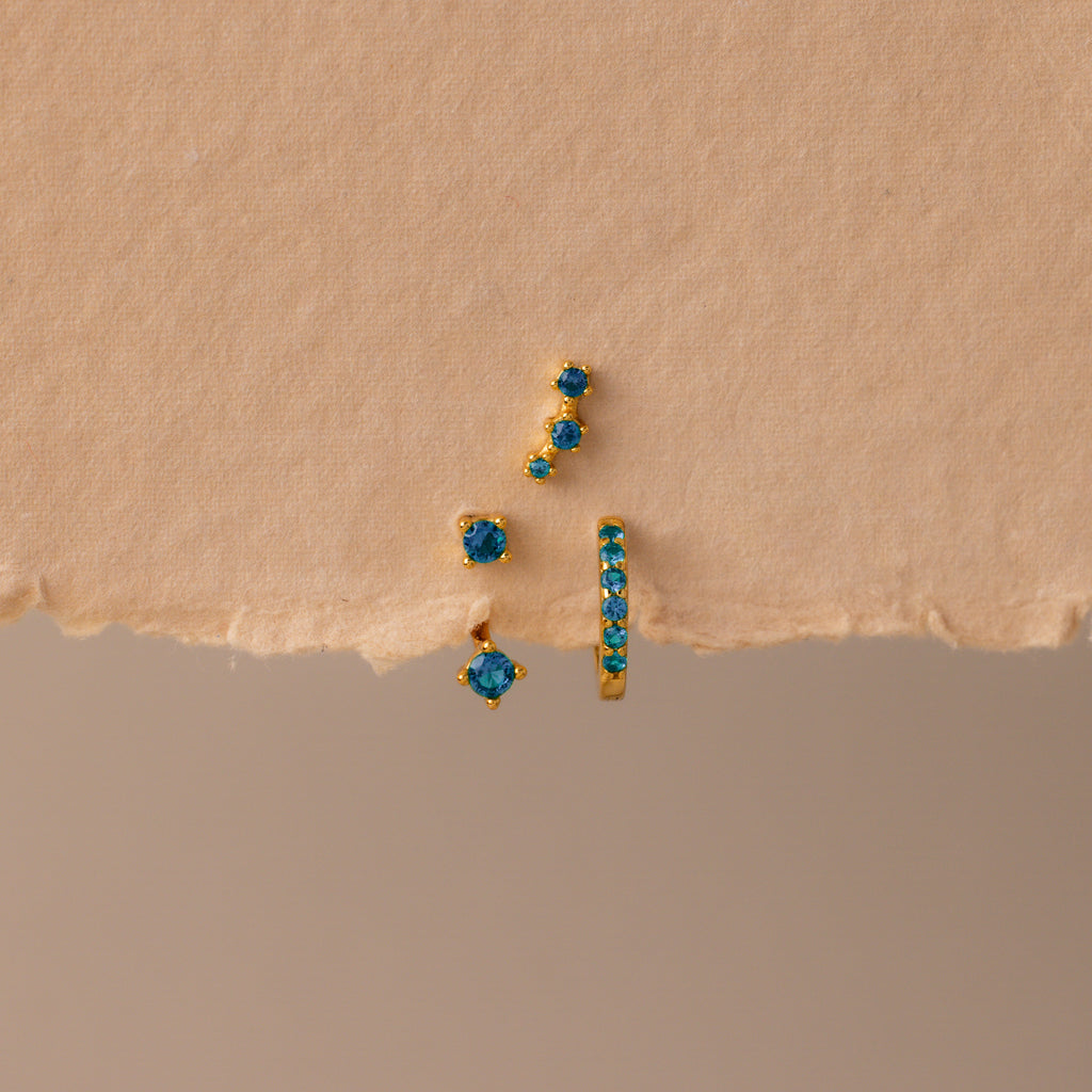 The Blue Zircon Earring Trio Set features three gold earrings with blue zircon stones—a small hoop and an elegant ear climber—displayed on textured beige paper, perfect for a unique birthstone earring set.