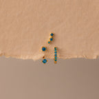 The Blue Zircon Earring Trio Set features three gold earrings with blue zircon stones—a small hoop and an elegant ear climber—displayed on textured beige paper, perfect for a unique birthstone earring set.