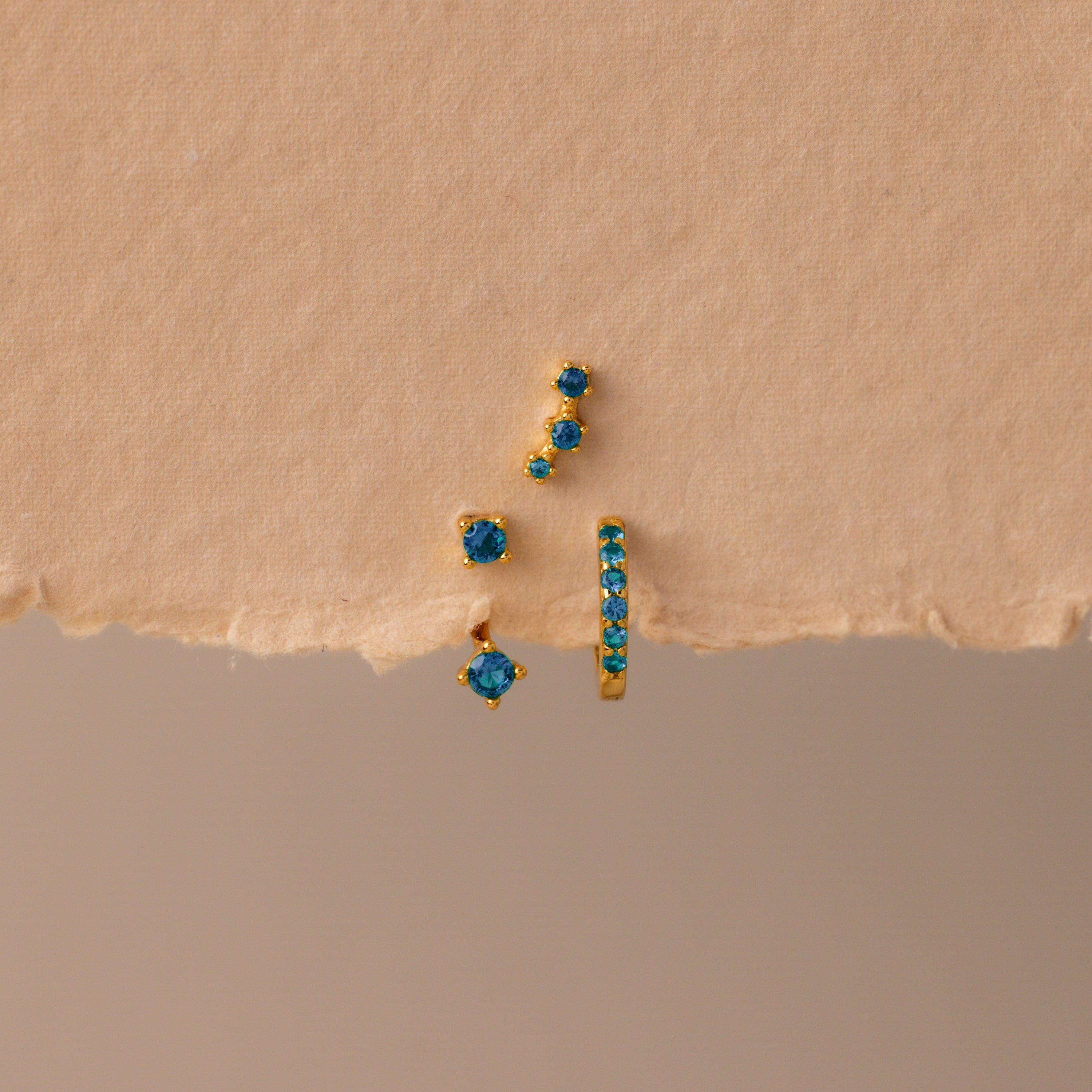 The Blue Zircon Earring Trio Set features three gold earrings with blue zircon stones—a small hoop and an elegant ear climber—displayed on textured beige paper, perfect for a unique birthstone earring set.