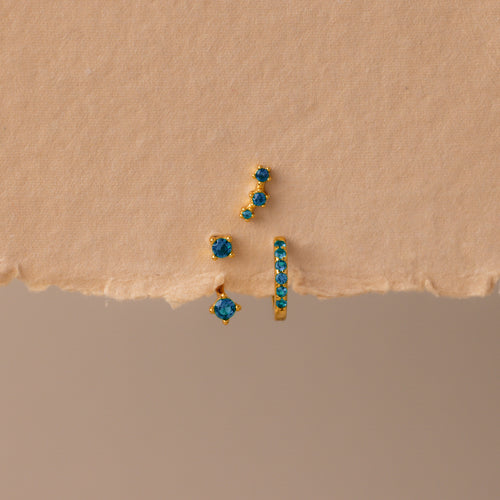 The Blue Zircon Earring Trio Set features three gold earrings with blue zircon stones—a small hoop and an elegant ear climber—displayed on textured beige paper, perfect for a unique birthstone earring set.