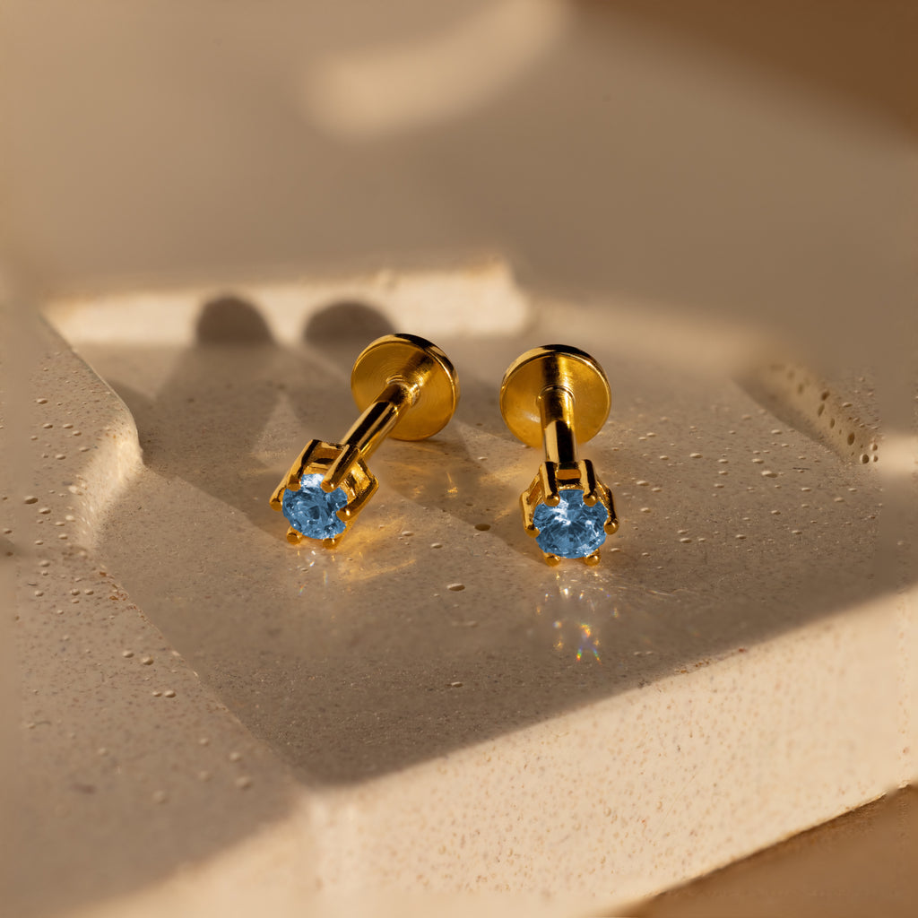 Blue Zircon Flatback Studs featuring blue gemstones are displayed on a textured, light-colored surface in gentle lighting.