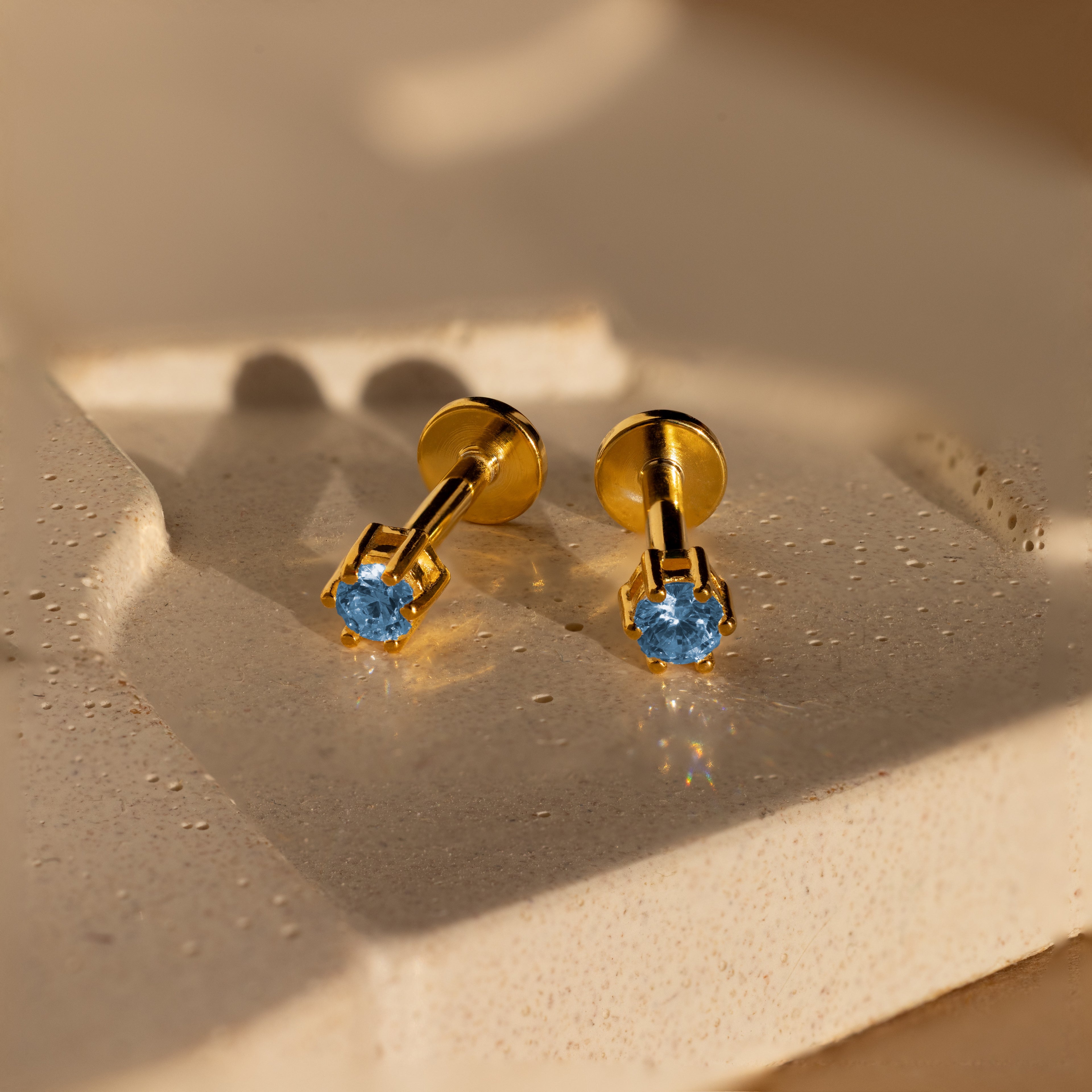 Blue Zircon Flatback Studs featuring blue gemstones are displayed on a textured, light-colored surface in gentle lighting.