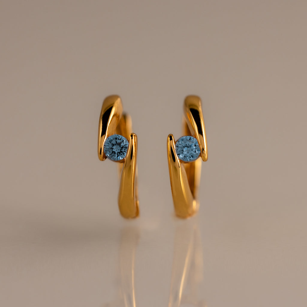 The Floating Blue Zircon Huggies feature modern curved gold hoops, each set with a single round blue zircon—perfect for lovers of personalized jewelry.