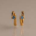 The Floating Blue Zircon Huggies feature modern curved gold hoops, each set with a single round blue zircon—perfect for lovers of personalized jewelry.
