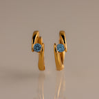 The Floating Blue Zircon Huggies feature modern curved gold hoops, each set with a single round blue zircon—perfect for lovers of personalized jewelry.