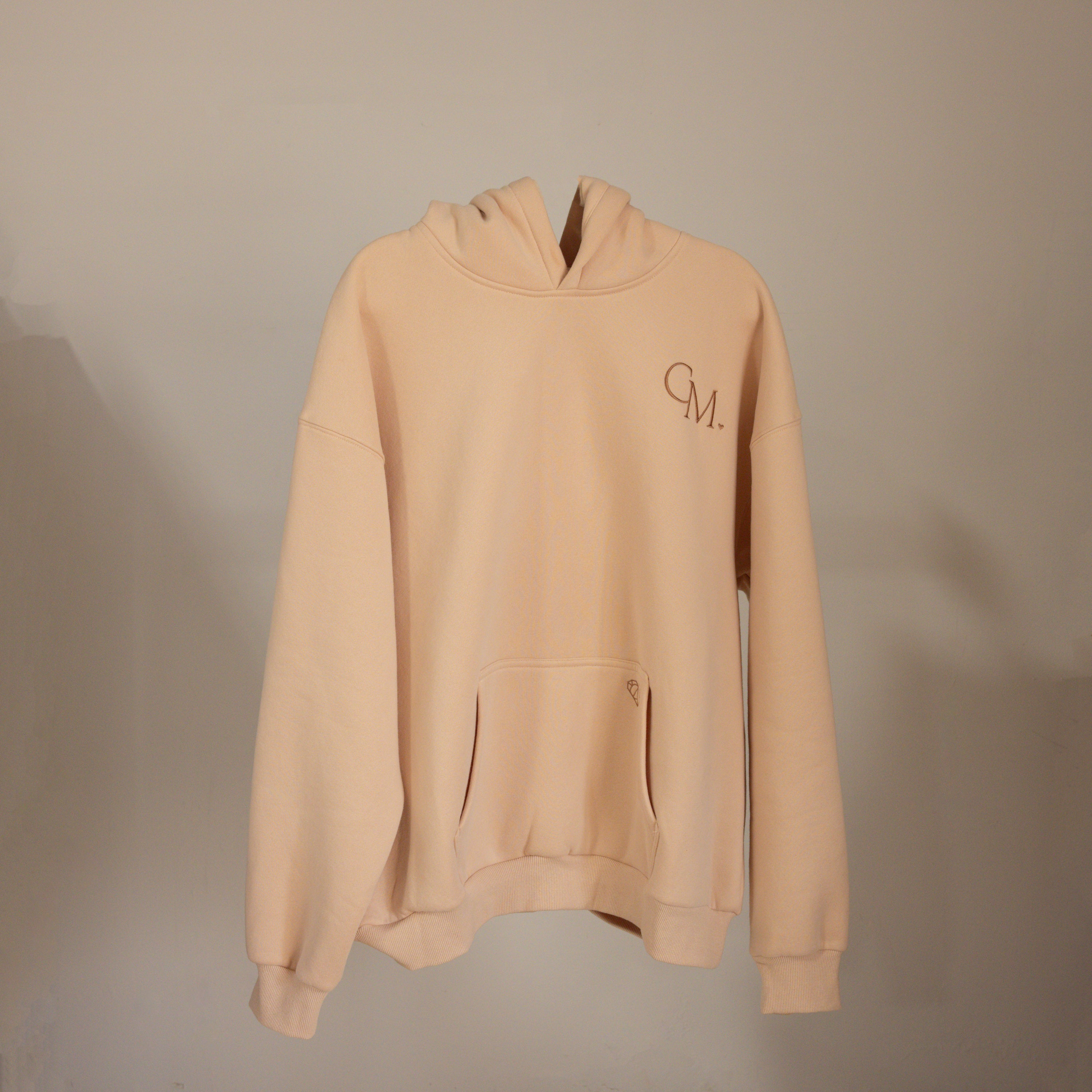 The Oversized CM Hoodie by Caitlyn Minimalist features a beige color, front pocket, and CM. logo on the left chest, displayed on a neutral background.