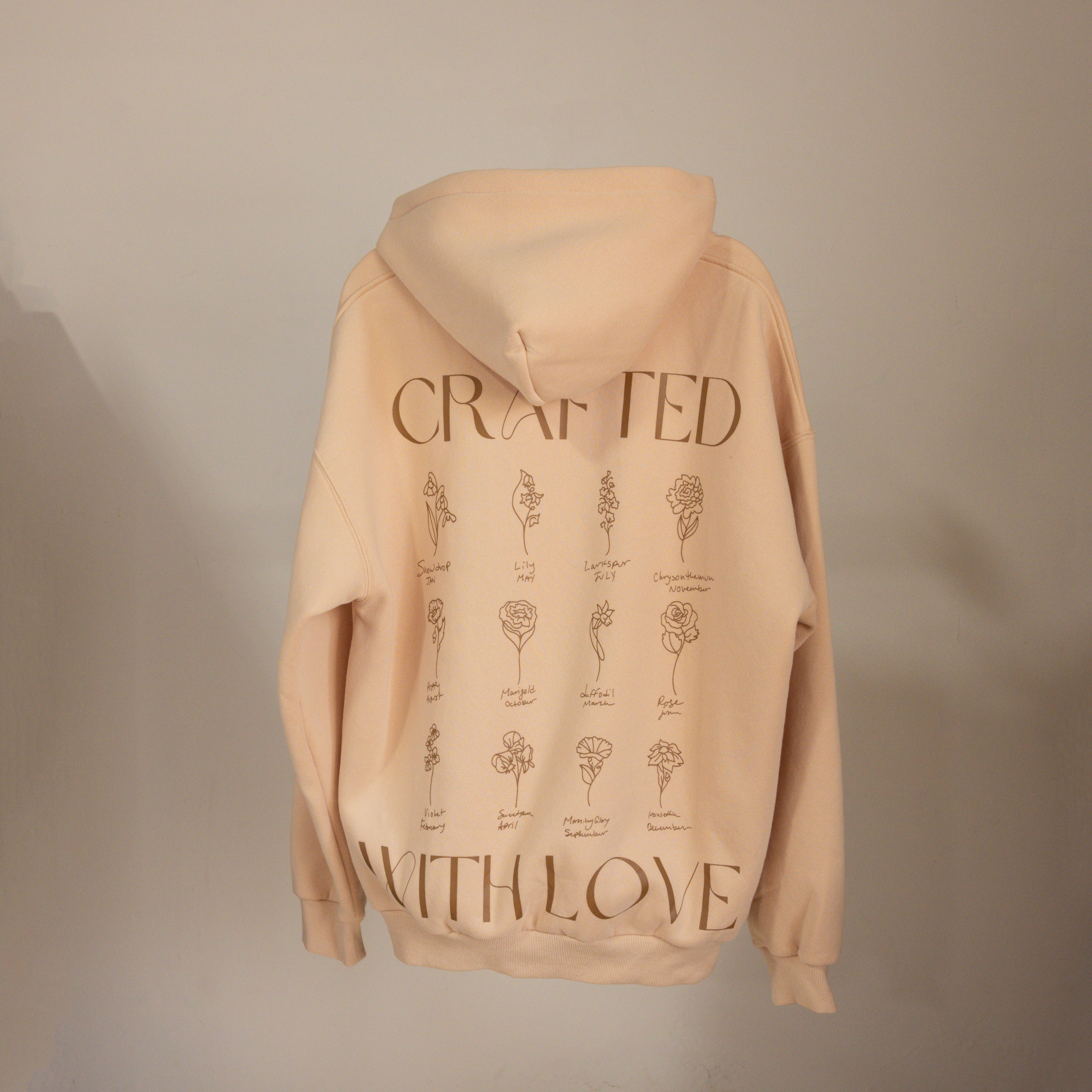 The Oversized CM Hoodie by Caitlyn Minimalist is a beige, cozy hoodie featuring hand-drawn flower line art with labeled names on the back.