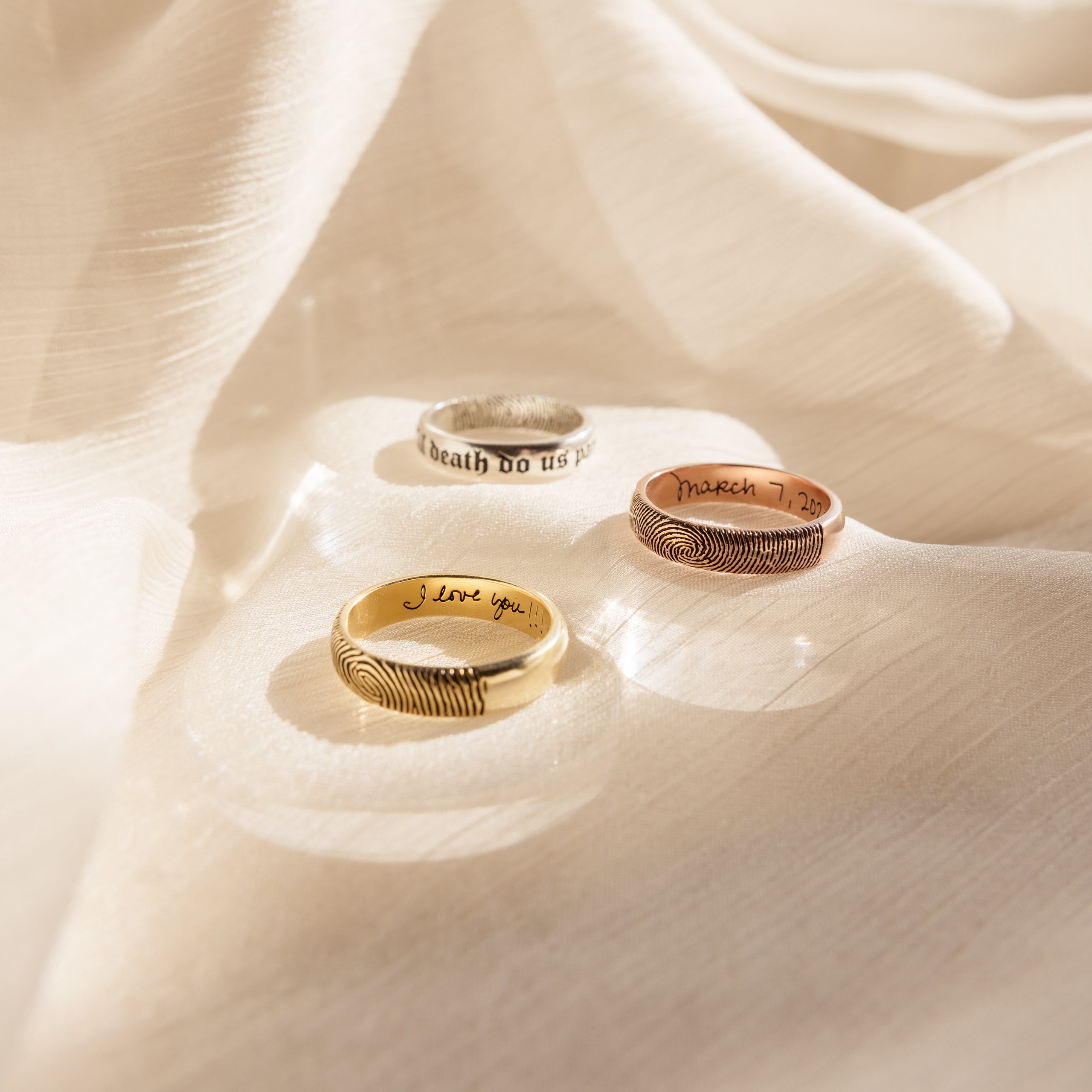 Three Half Round Fingerprint Rings, each engraved with a unique fingerprint design, rest on soft, light-colored fabric in warm sunlight, highlighting the personalized style of the Half Round Fingerprint Ring.