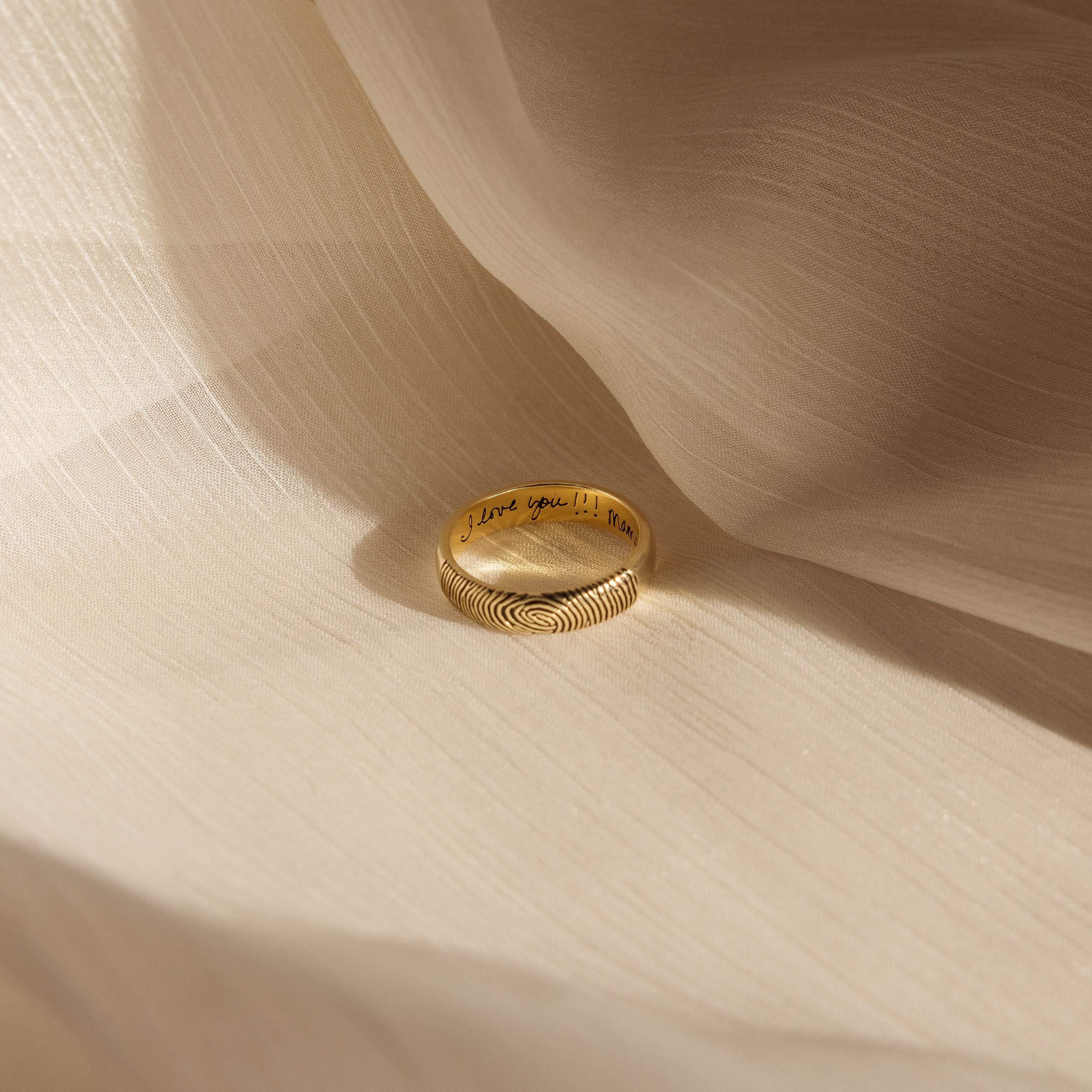 The Half Round Fingerprint Ring, engraved with "I love you!!", rests on soft cream fabric—this personalized piece of jewelry is uniquely crafted for you.