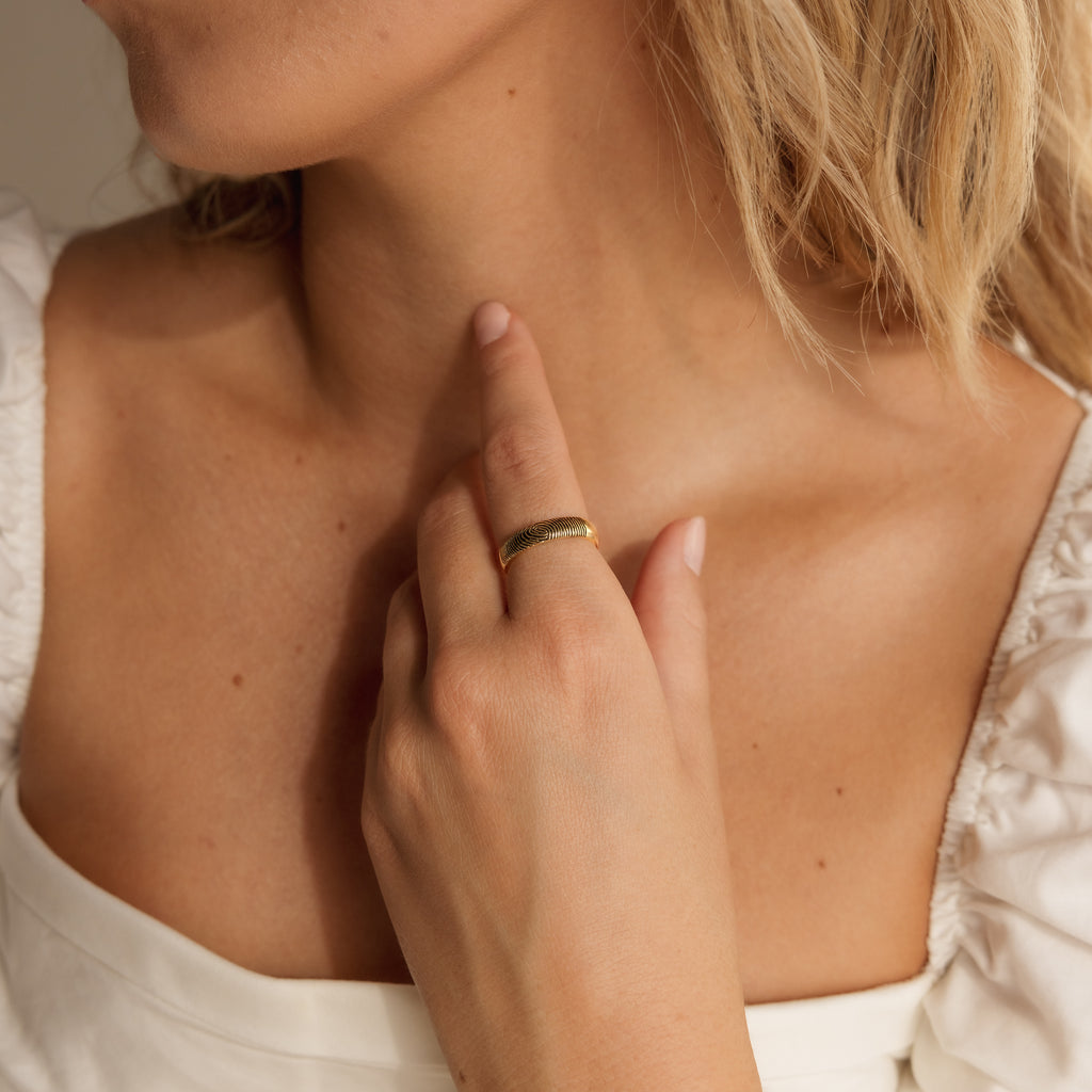 A woman in a white top touches her neck, highlighting the gold Half Round Fingerprint Ring on her finger—a unique and personalized jewelry piece.