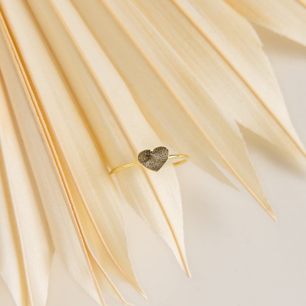 The Fingerprint Heart Ring features a textured heart design, shown on cream-colored dried palm leaves—ideal for anyone looking for elegant, personalized jewelry.