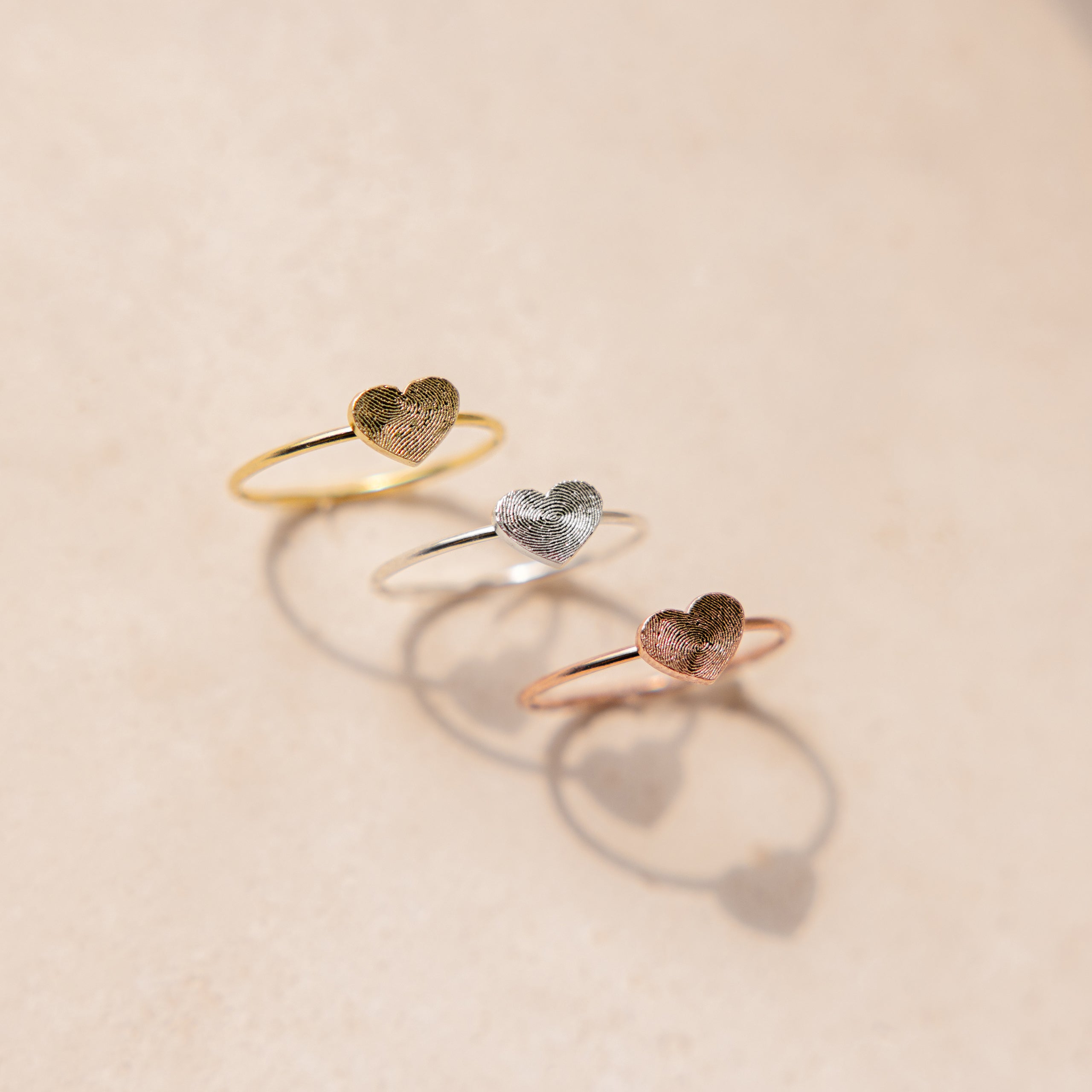 The Fingerprint Heart Ring features delicate heart designs in gold, silver, and rose gold on a beige background—ideal as custom jewelry or unique personalized gifts.