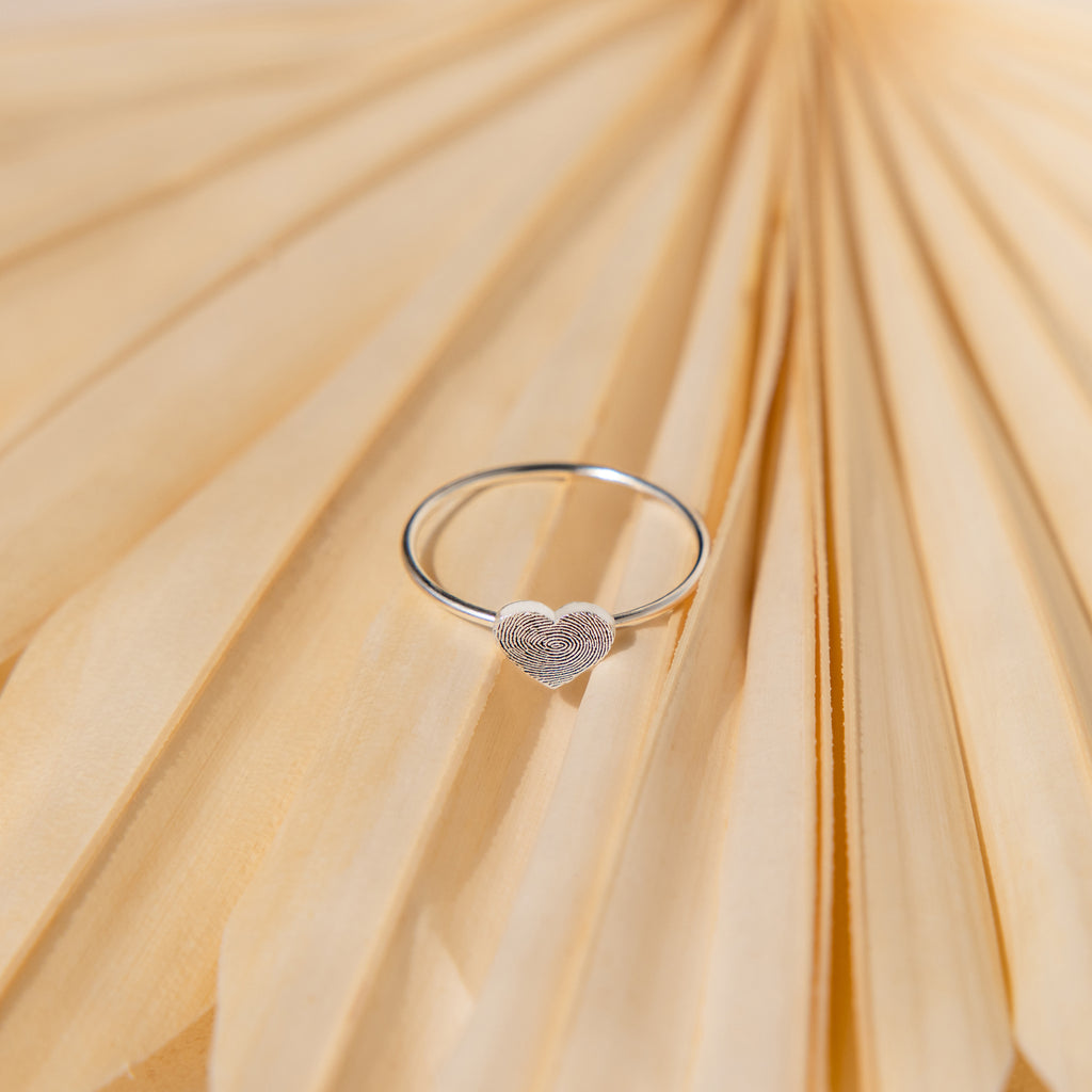 The Fingerprint Heart Ring, featuring a heart-shaped design, is elegantly displayed on a light beige fan-shaped surface—an exquisite piece of personalized jewelry.
