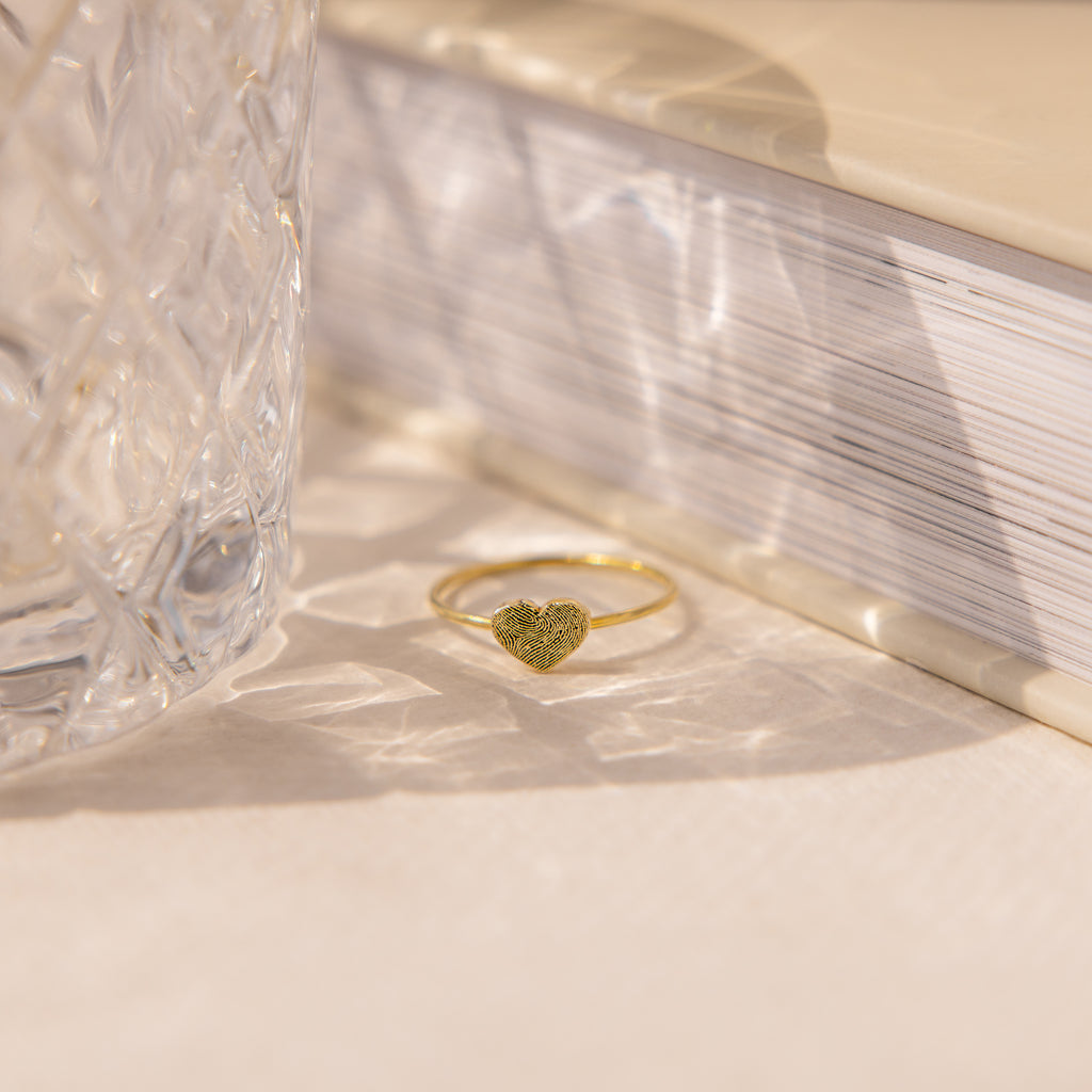 The Fingerprint Heart Ring, crafted in gold, lies on a light surface next to a crystal glass and closed book—a custom piece that captures your unique love and memories.