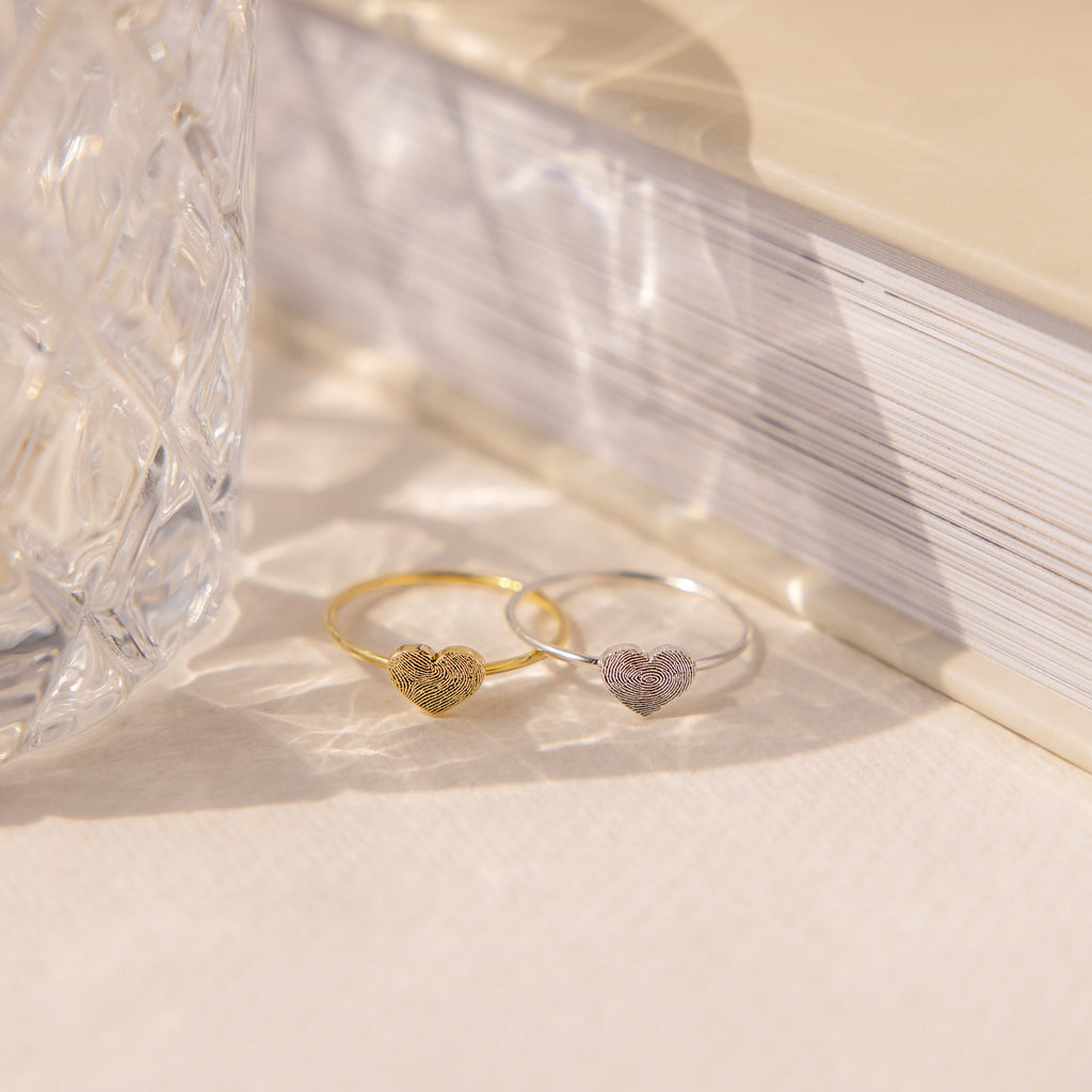 A gold and a silver heart-shaped ring sit by textured glass and a closed book—showcasing custom jewelry such as the Fingerprint Heart Ring.
