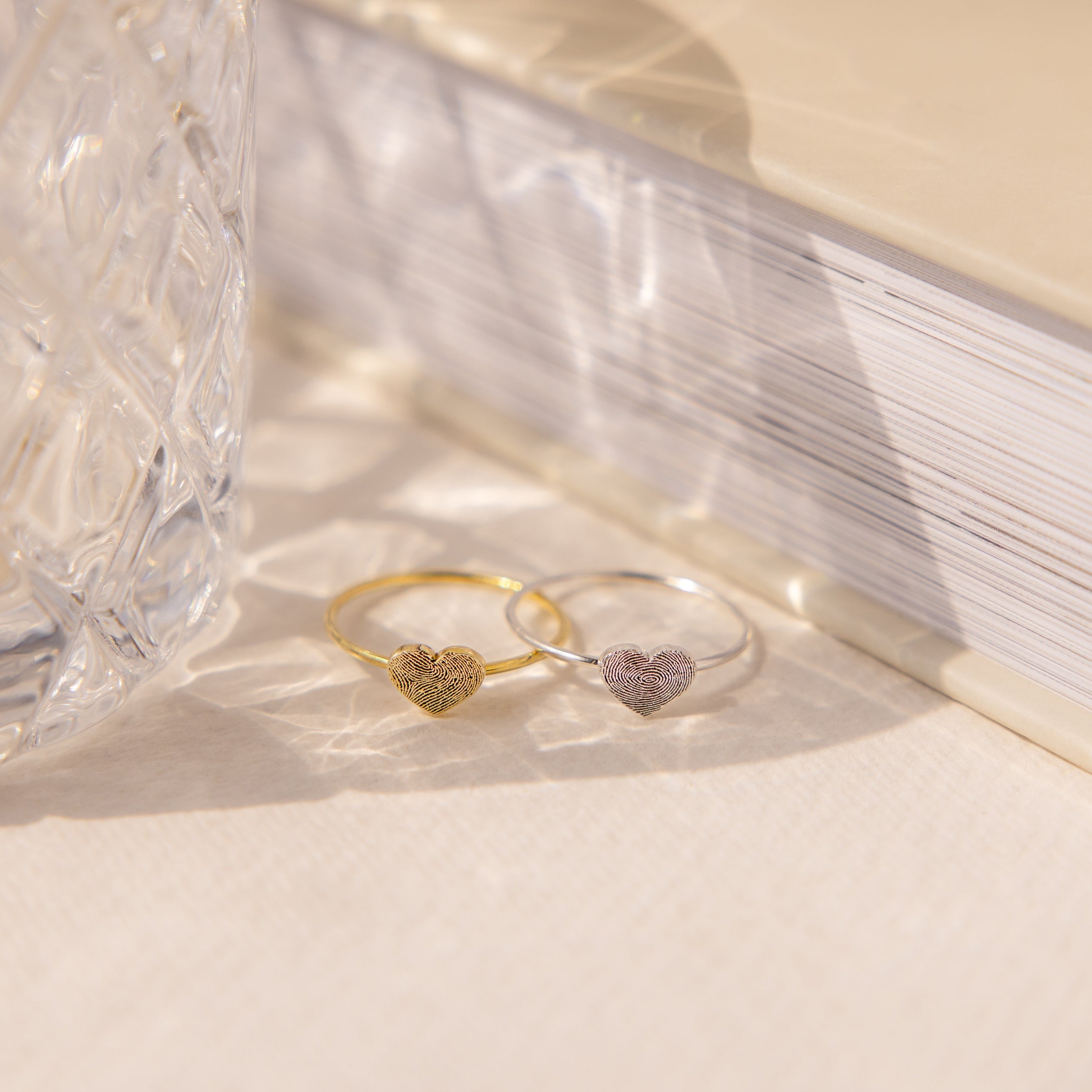 A gold and a silver heart-shaped ring sit by textured glass and a closed book—showcasing custom jewelry such as the Fingerprint Heart Ring.