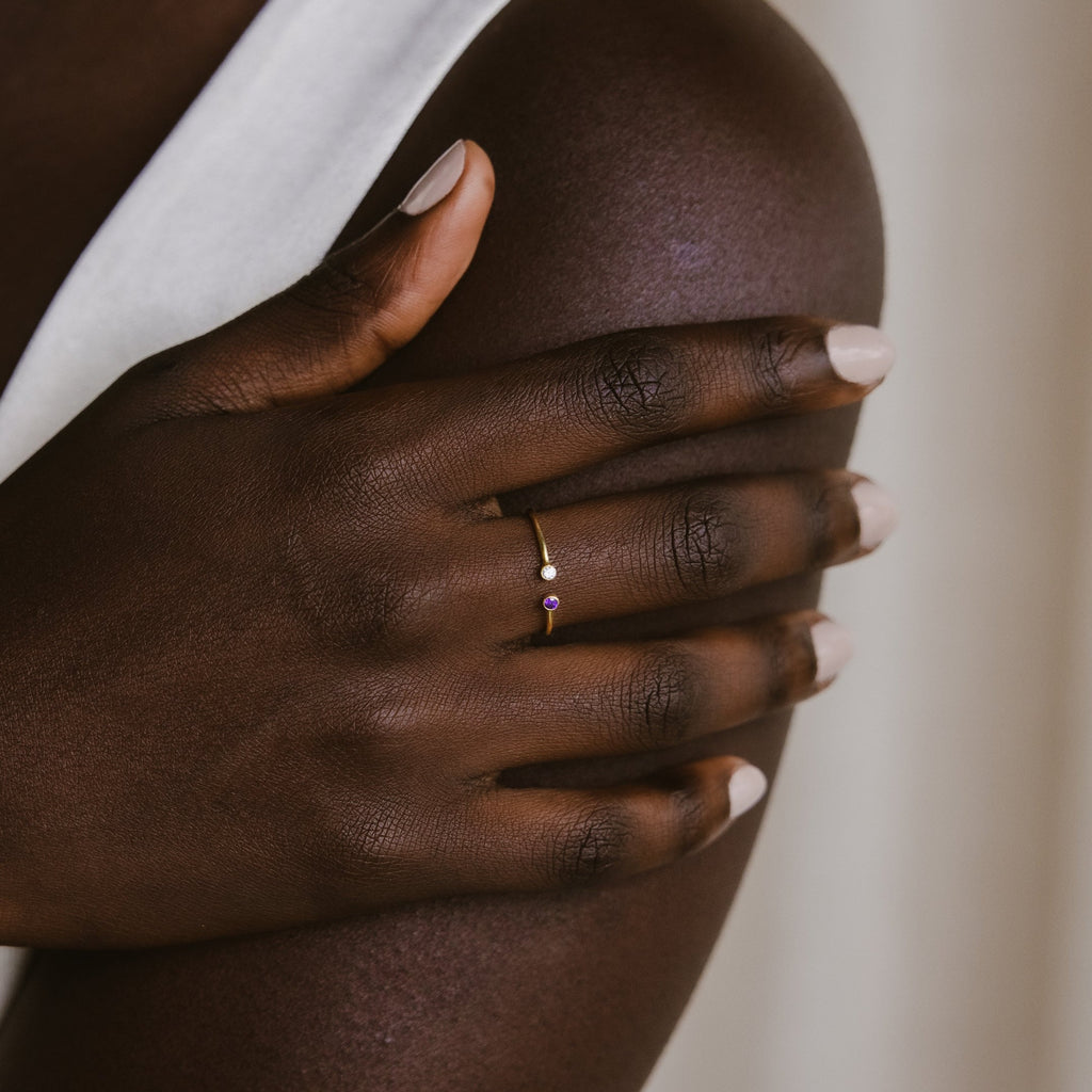 A hand adorned with the Tiny Dual Birthstone Ring holds an upper arm, nails painted light beige, and wears a sleeveless white top.