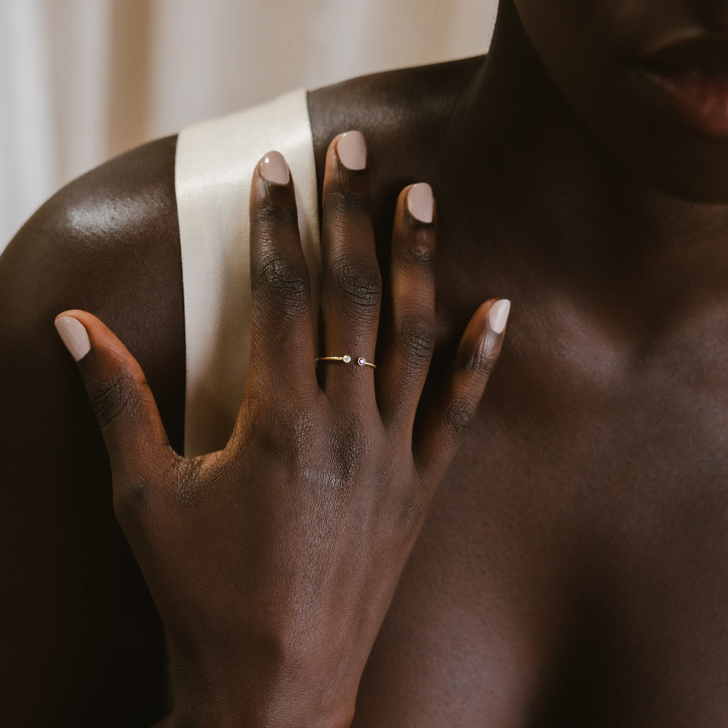 A hand with light pink nails rests on a shoulder, adorned with the Tiny Dual Birthstone Ring—a delicate gold band set with two subtle gemstones.