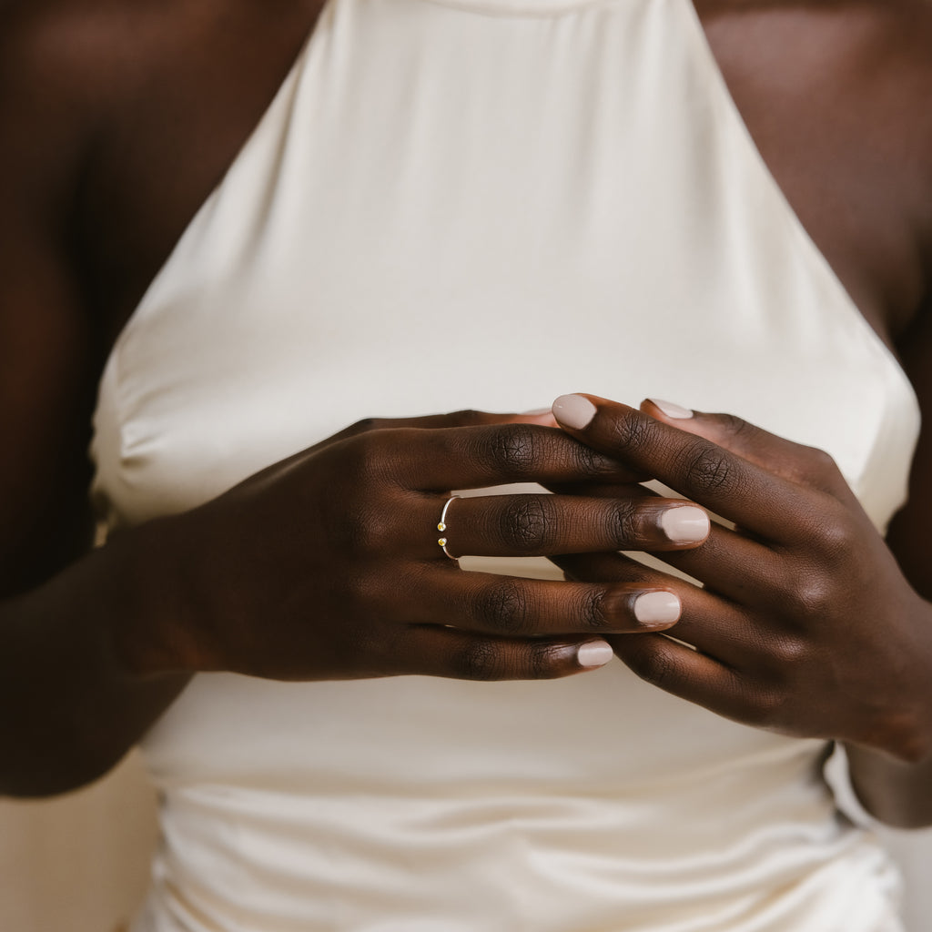 A woman in a white dress gently clasps her hands, showcasing the Tiny Dual Birthstone Ring on her finger.