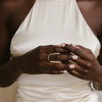 A woman in a white dress gently clasps her hands, showcasing the Tiny Dual Birthstone Ring on her finger.