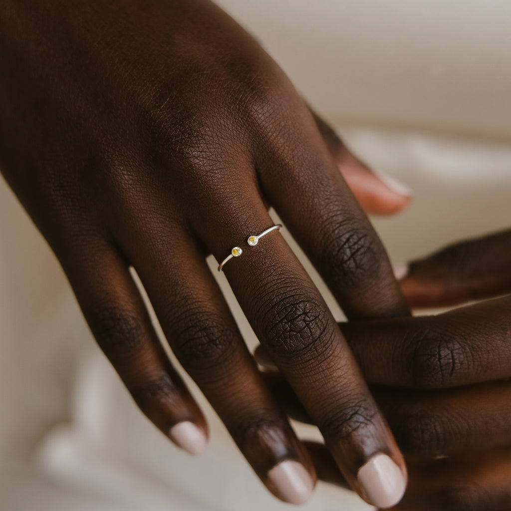 A hand wears the Tiny Dual Birthstone Ring—a delicate silver band with two small stones—on the ring finger.