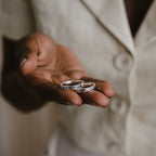 A hand holds two silver Mothers Birthstone Rings, each engraved and personalized as a keepsake; the person wears a light-colored button-up shirt.