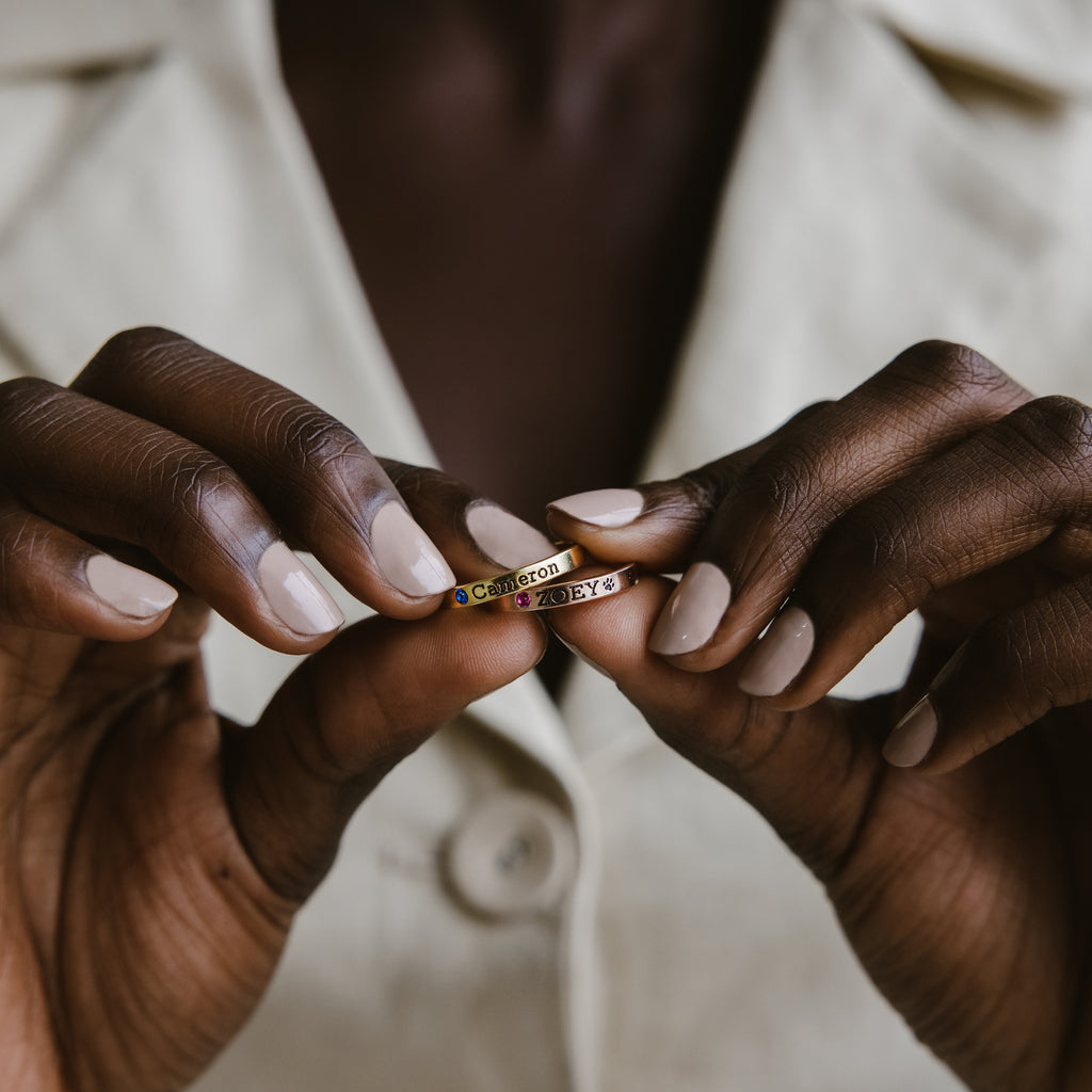 A person wearing a beige jacket holds a Stacking Name Band— a personalized gold ring engraved with "Cameroon" and unique symbols.