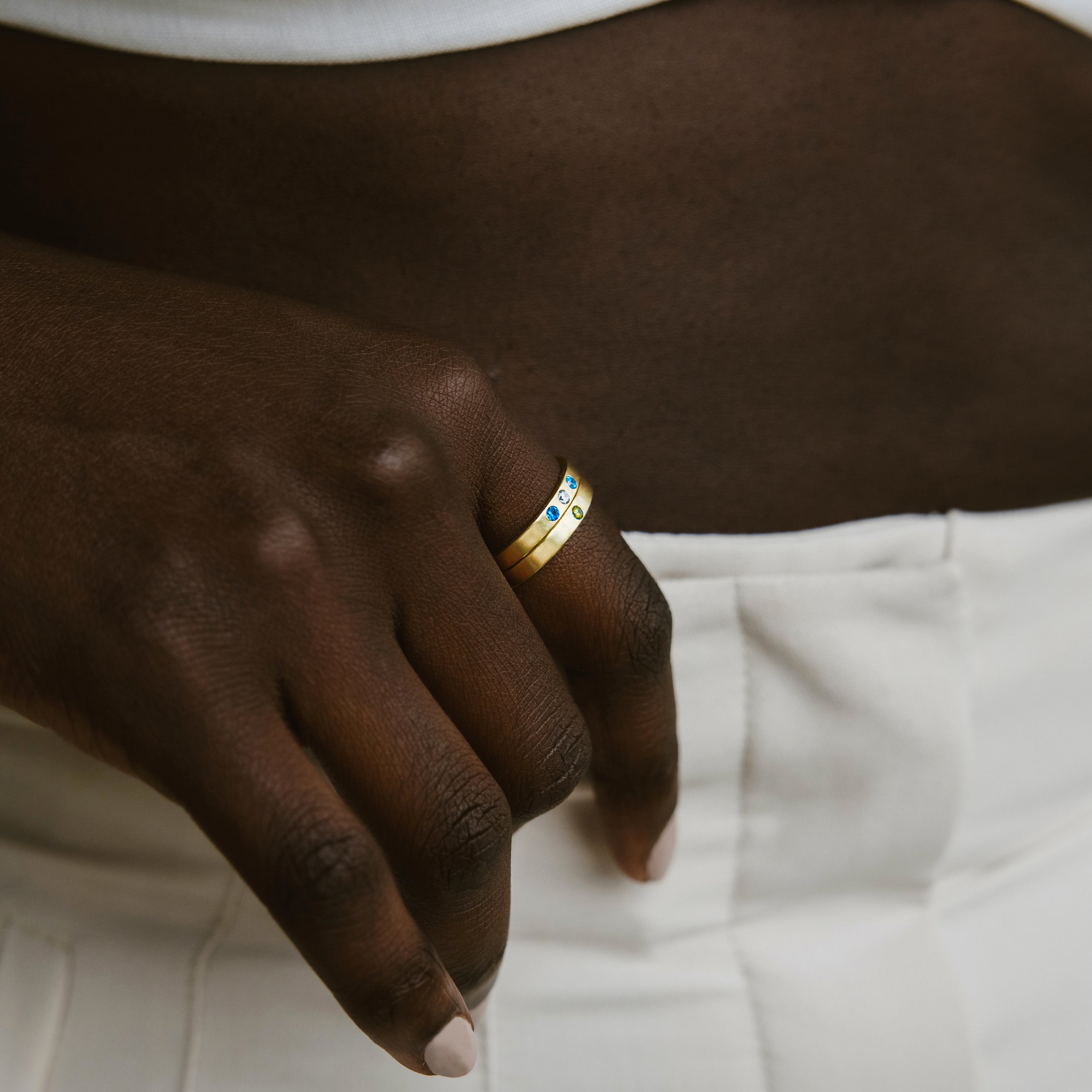 A hand adorned with the Mothers Birthstone Ring rests on white pants, its gold bands and personalized birthstones shimmering elegantly against a brown midriff—a timeless keepsake for any occasion.