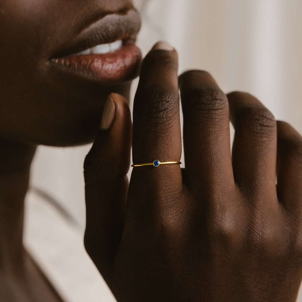 A close-up shows a hand wearing the Tiny Birthstone Ring, which features a small blue gemstone and delicate detailing, held near the person's lips.