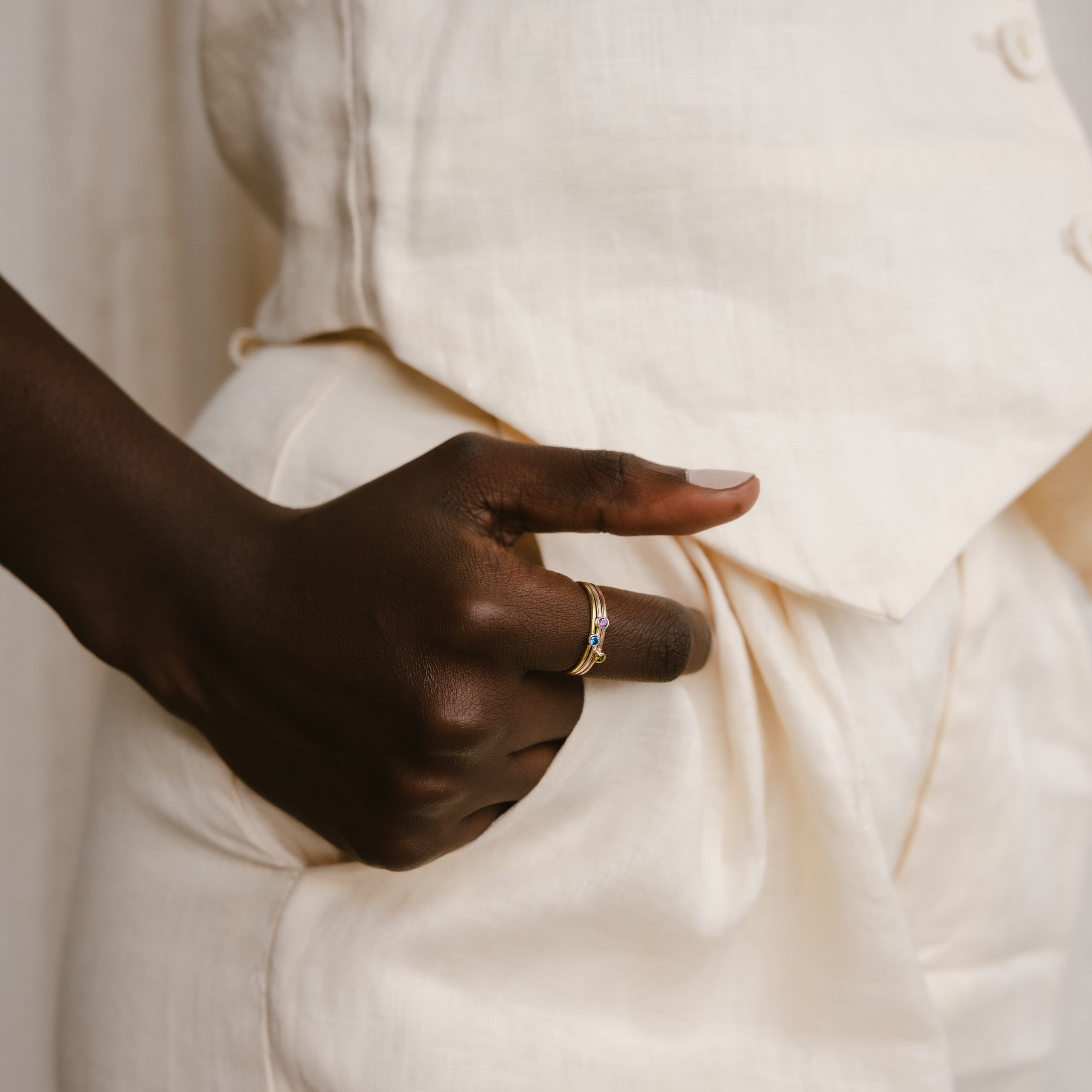 A hand wearing the Tiny Birthstone Ring rests on the pocket of cream pants with a matching top, adding a subtle touch of birthstone color to the outfit.