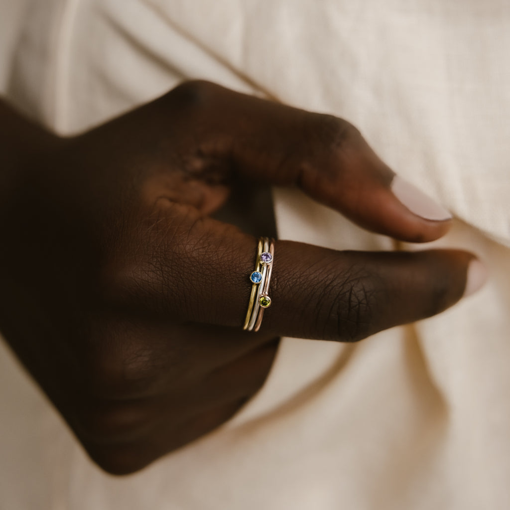 A hand wearing two Tiny Birthstone Rings, each adorned with gemstone accents in birthstone hues, is set against a cream-colored fabric backdrop.