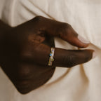 A hand wearing two Tiny Birthstone Rings, each adorned with gemstone accents in birthstone hues, is set against a cream-colored fabric backdrop.