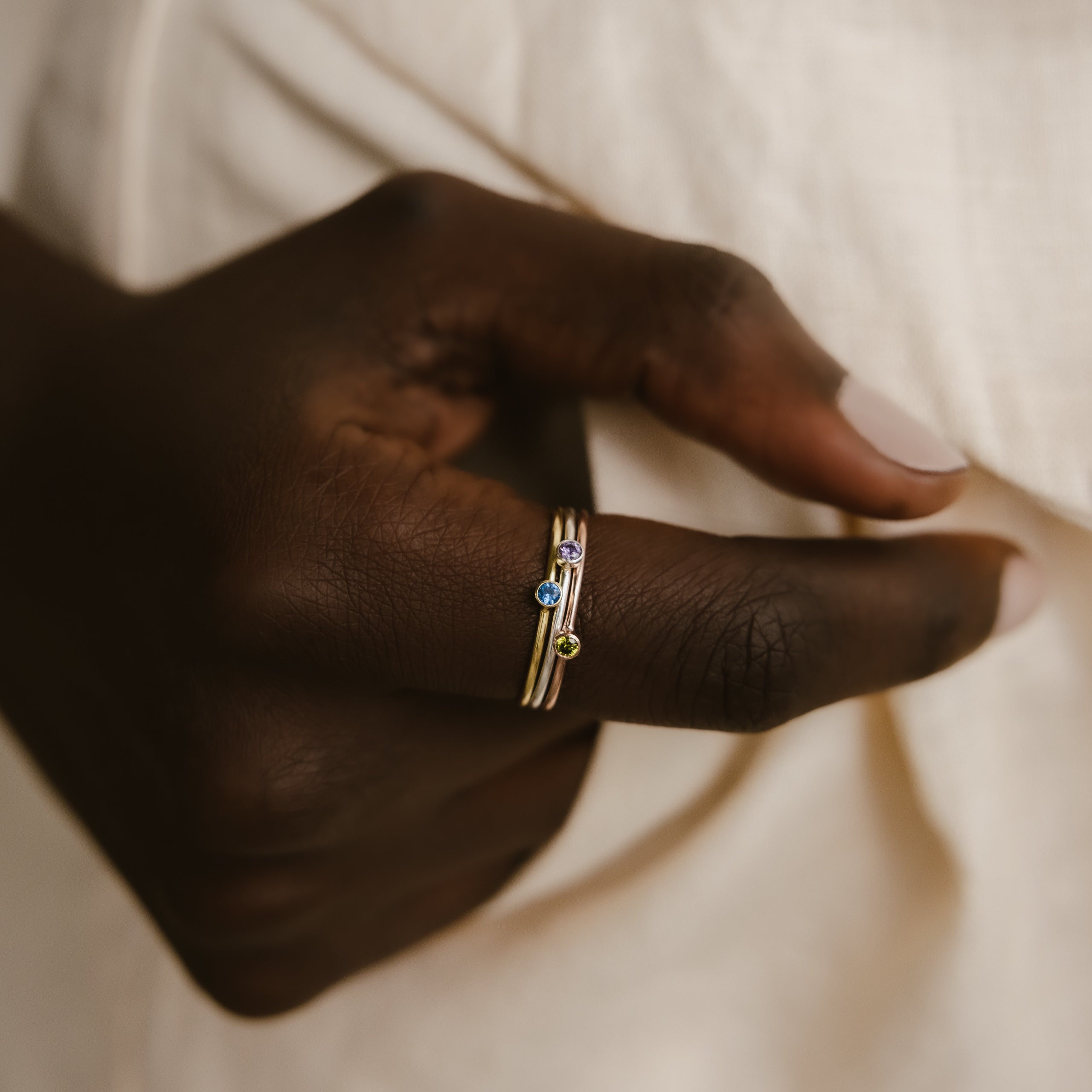 A hand wearing two Tiny Birthstone Rings, each adorned with gemstone accents in birthstone hues, is set against a cream-colored fabric backdrop.